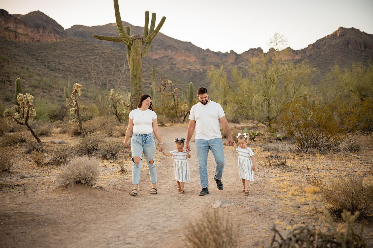 Family of four walks hand-in-hand on a desert trail surrounded by saguaro cacti and mountain scenery.