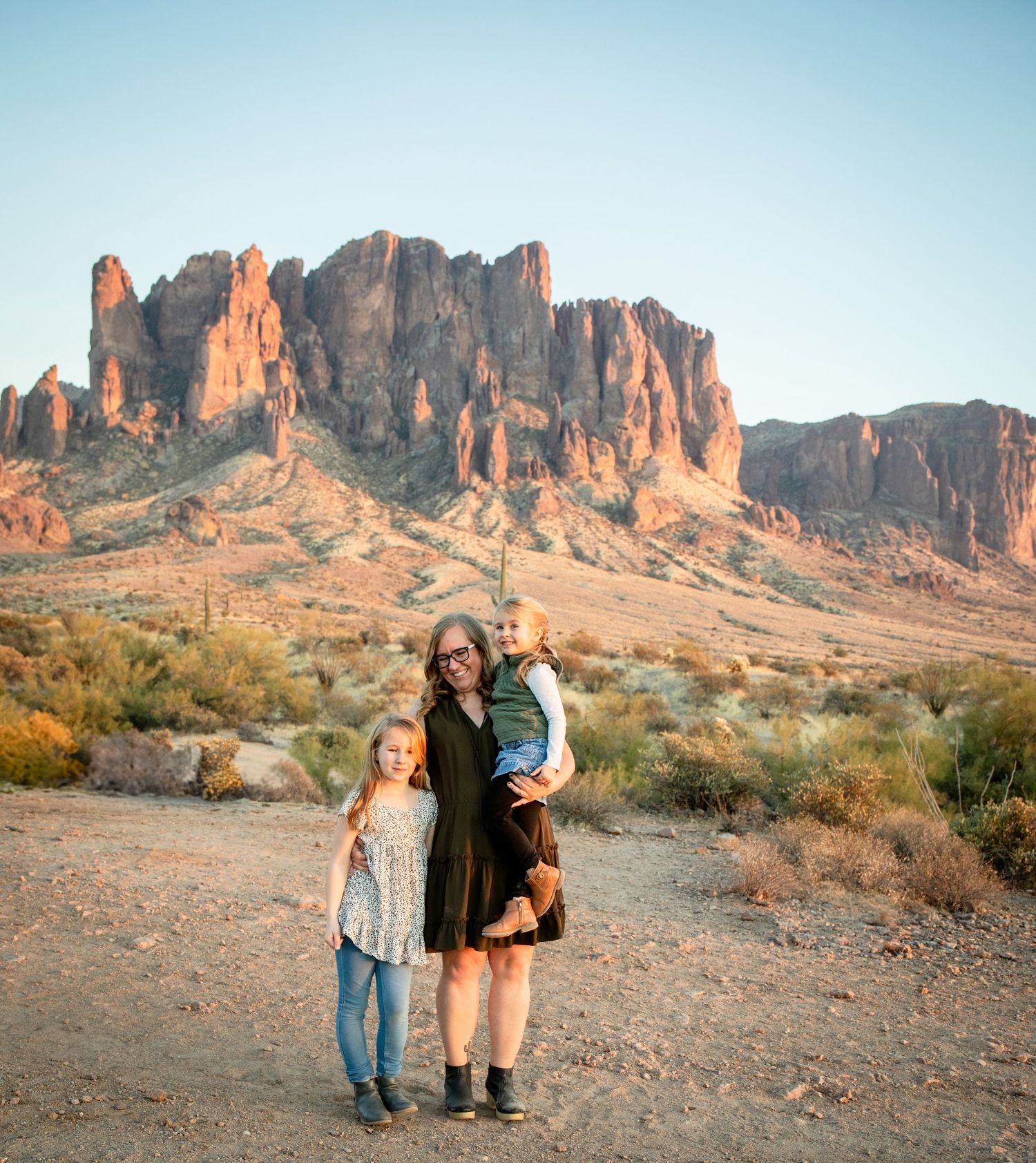 A family stands together on a desert trail with the majestic Superstition Mountains glowing orange at sunset in Arizona.