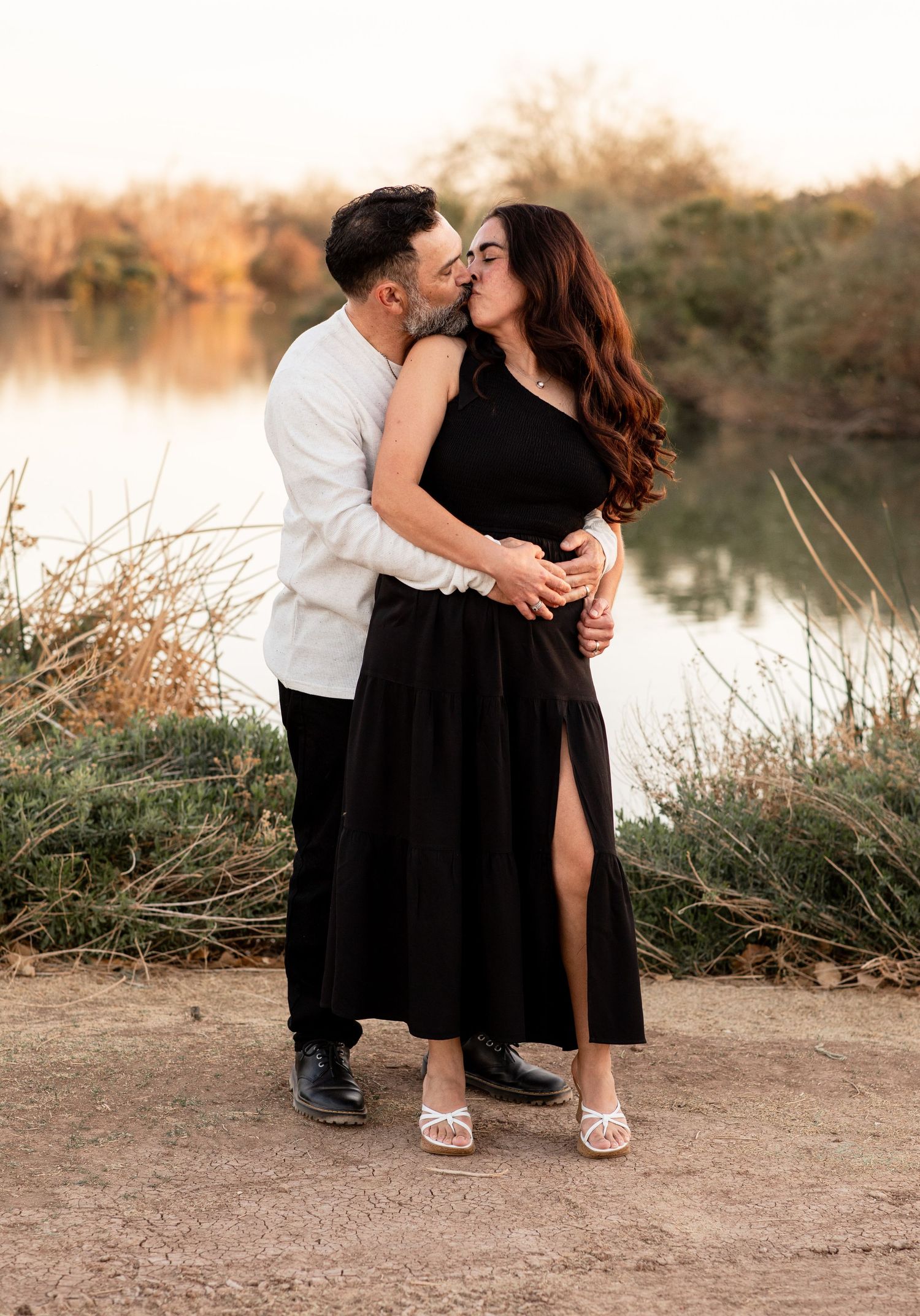 A romantic couple in black and white outfits embraces by a tranquil lake during sunset in a series of intimate poses.