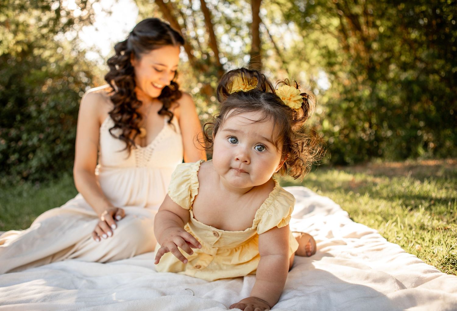 A young child in a gold crown sits on a white blanket in a sunlit outdoor setting during a photo session at The Meadow in Mesa.