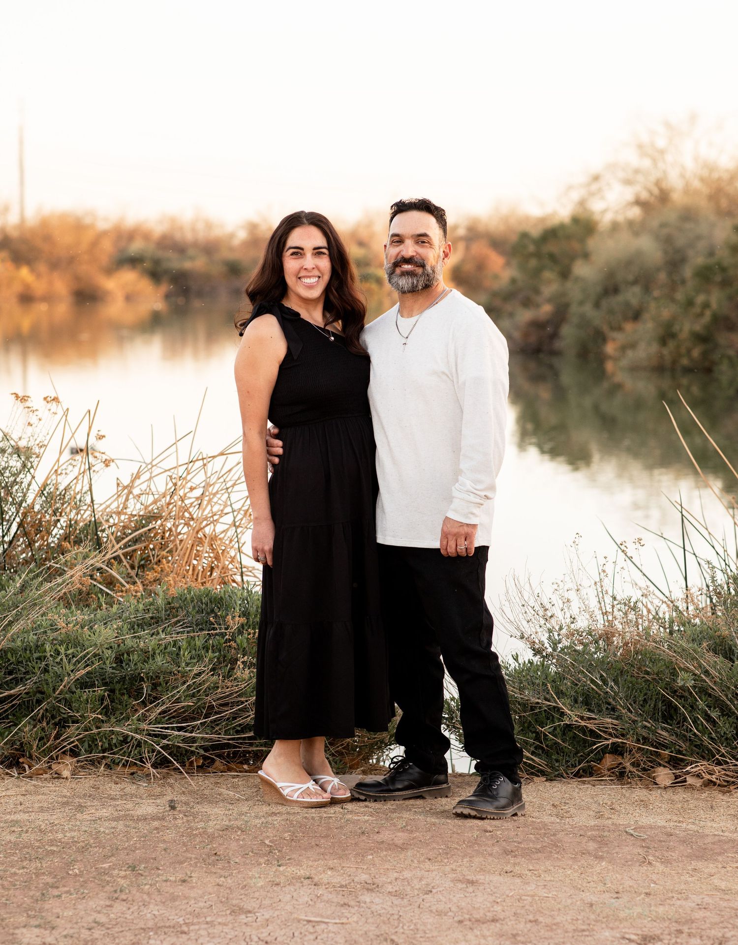 Couple standing together at sunset by a lake with tall grass and natural desert landscape in the background.