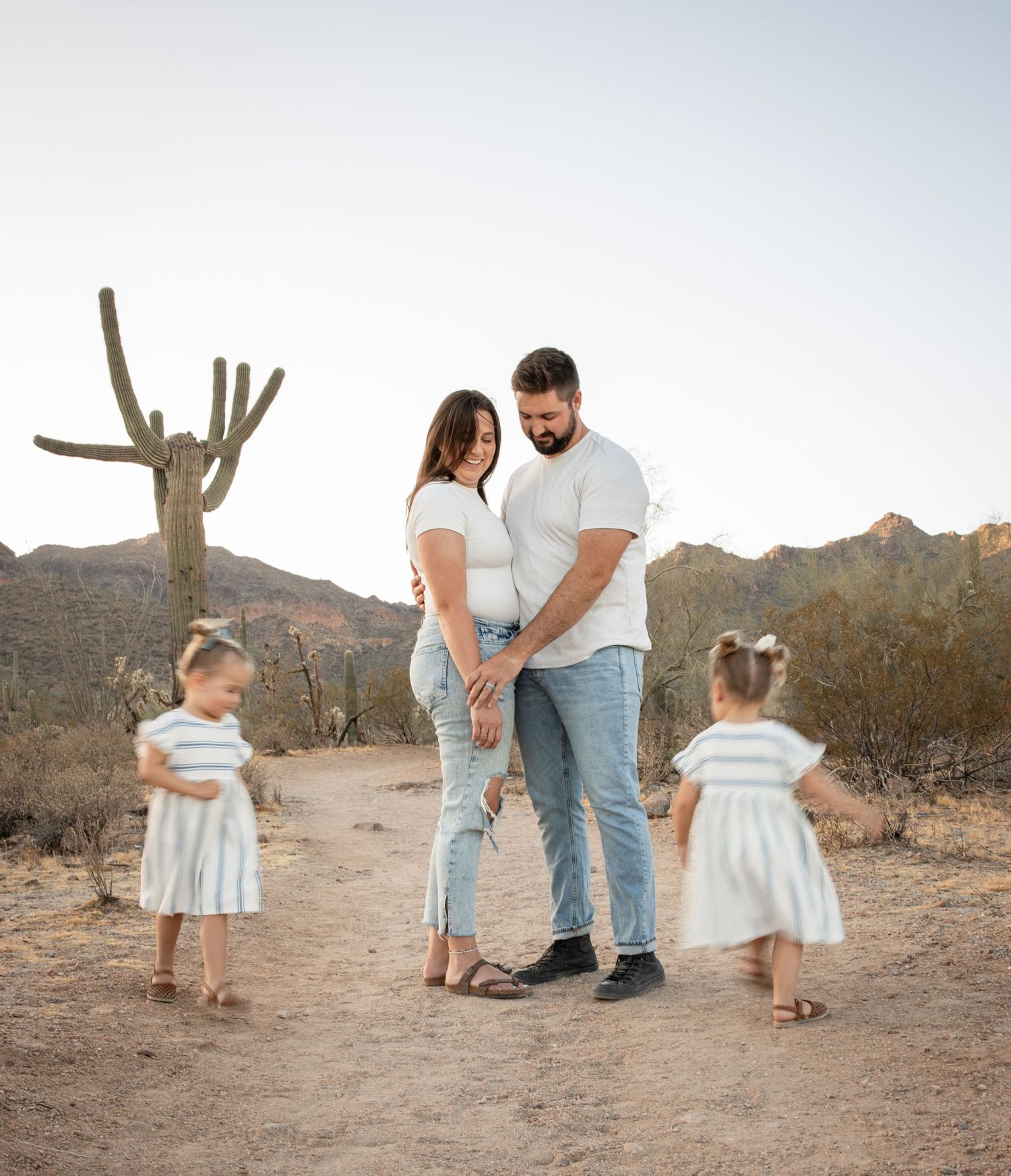 Pregnant couple in white tops holding hands in Arizona desert with two young daughters in striped dresses and saguaro cactus.