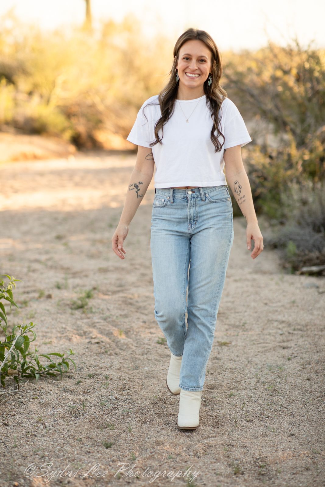 Person in light blue jeans and white t-shirt walks on desert trail during golden hour sunset.