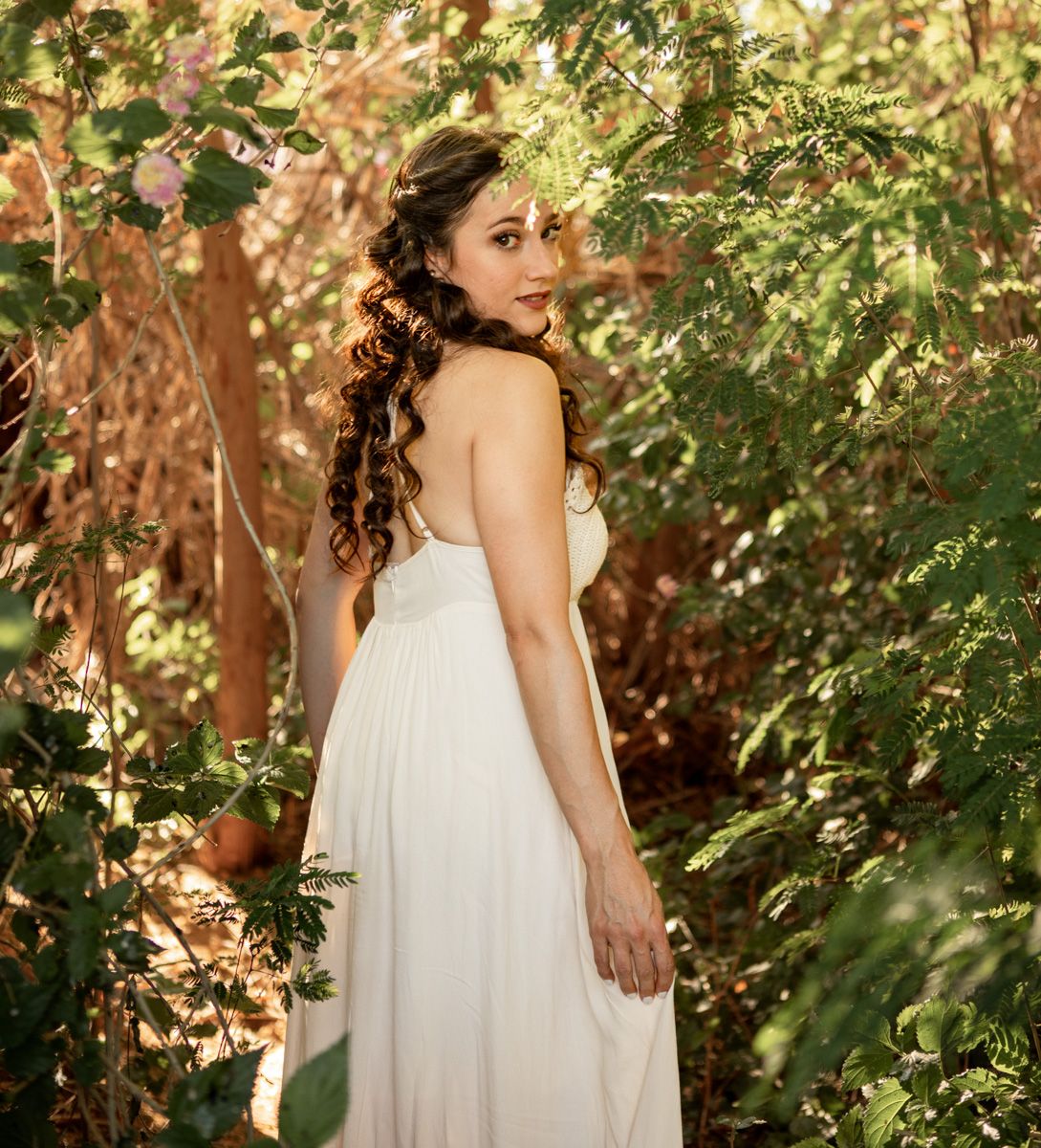 A woman in a flowing white dress poses gracefully in a garden setting with dappled sunlight and green foliage at The Meadow in Mesa, AZ.