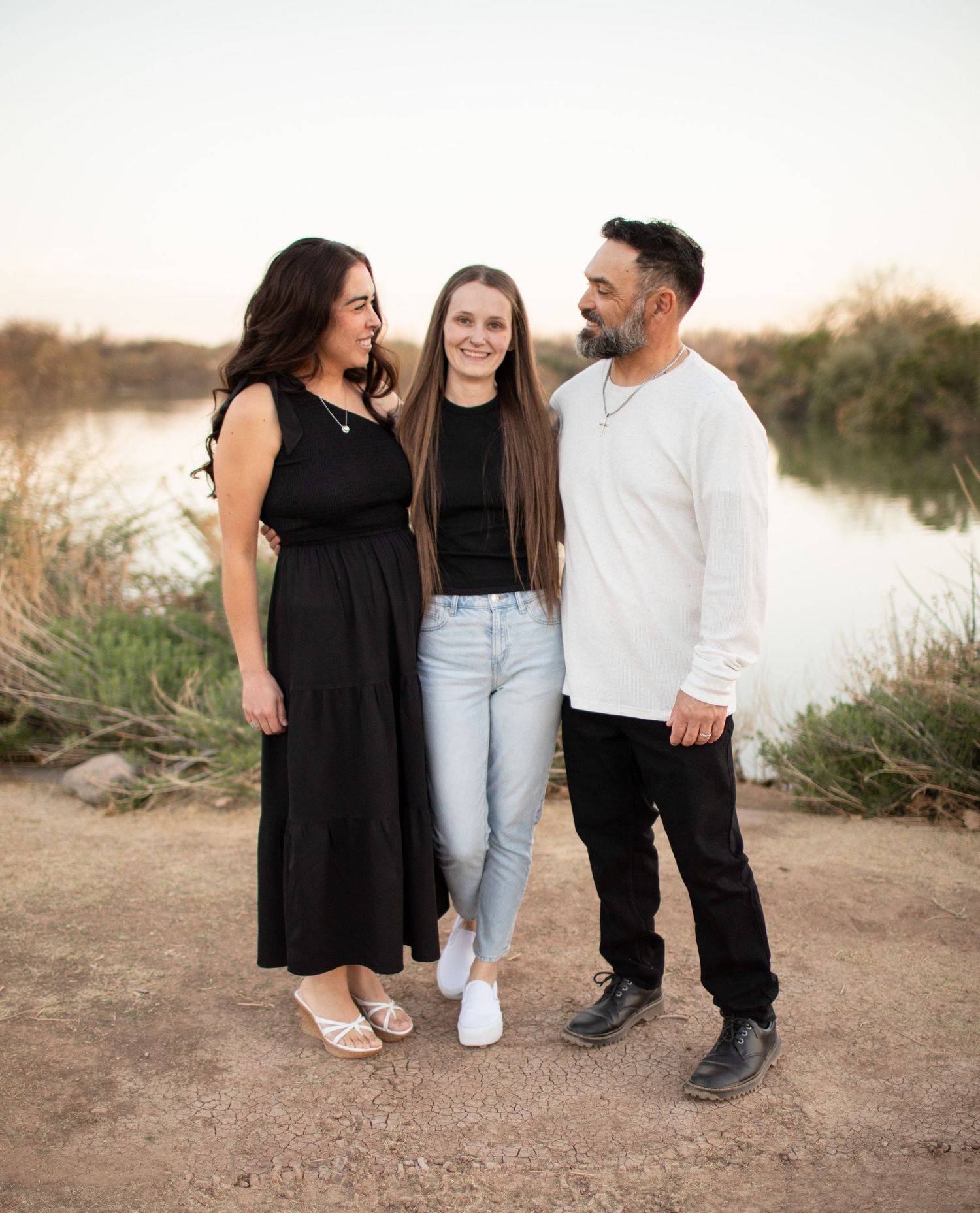 A family in casual attire stands together on a dirt path near a lake during golden hour.