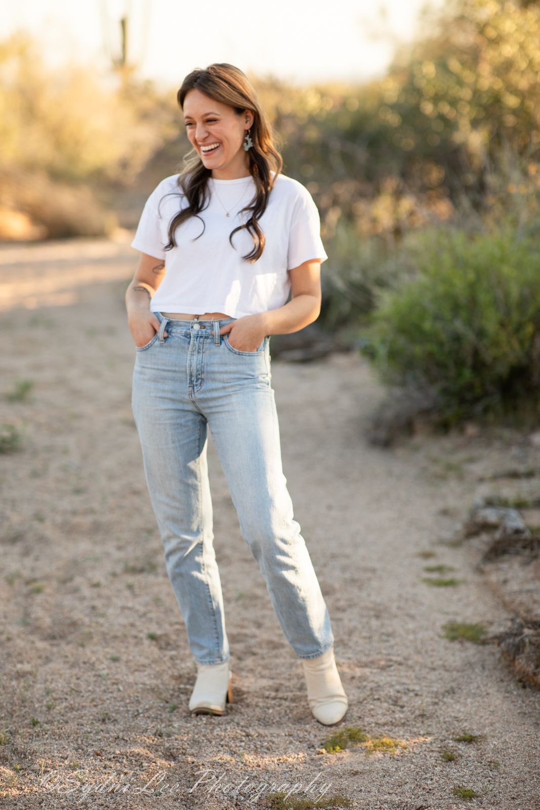 Person wearing casual white t-shirt and light jeans poses on dirt path with desert vegetation.