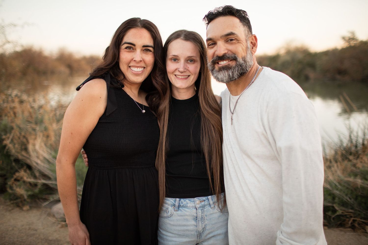 Three people wearing black and white clothing pose together outdoors during golden hour at sunset.