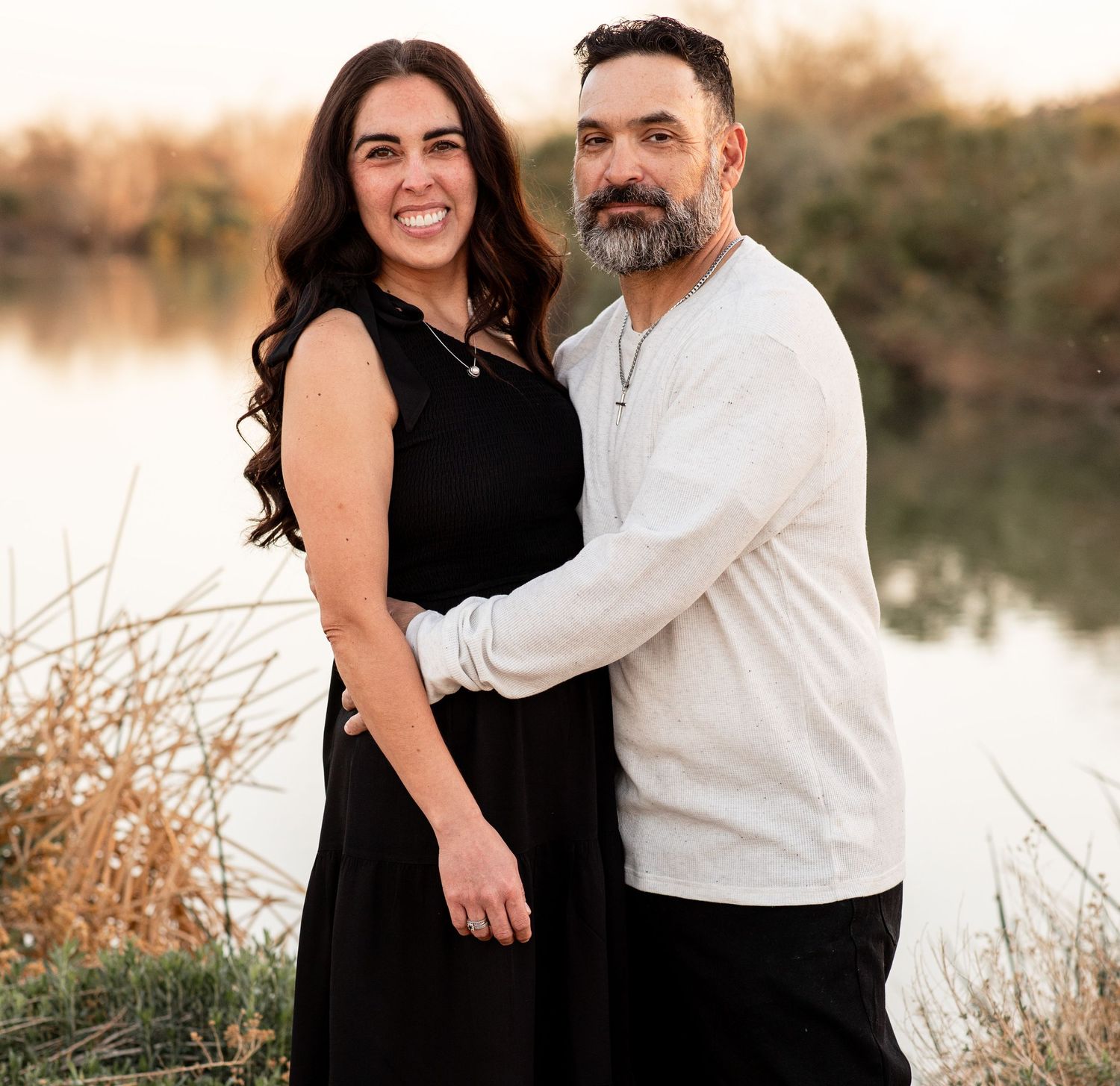 A couple poses together outdoors in black and white outfits during a sunset photoshoot by a lake with tall grasses.