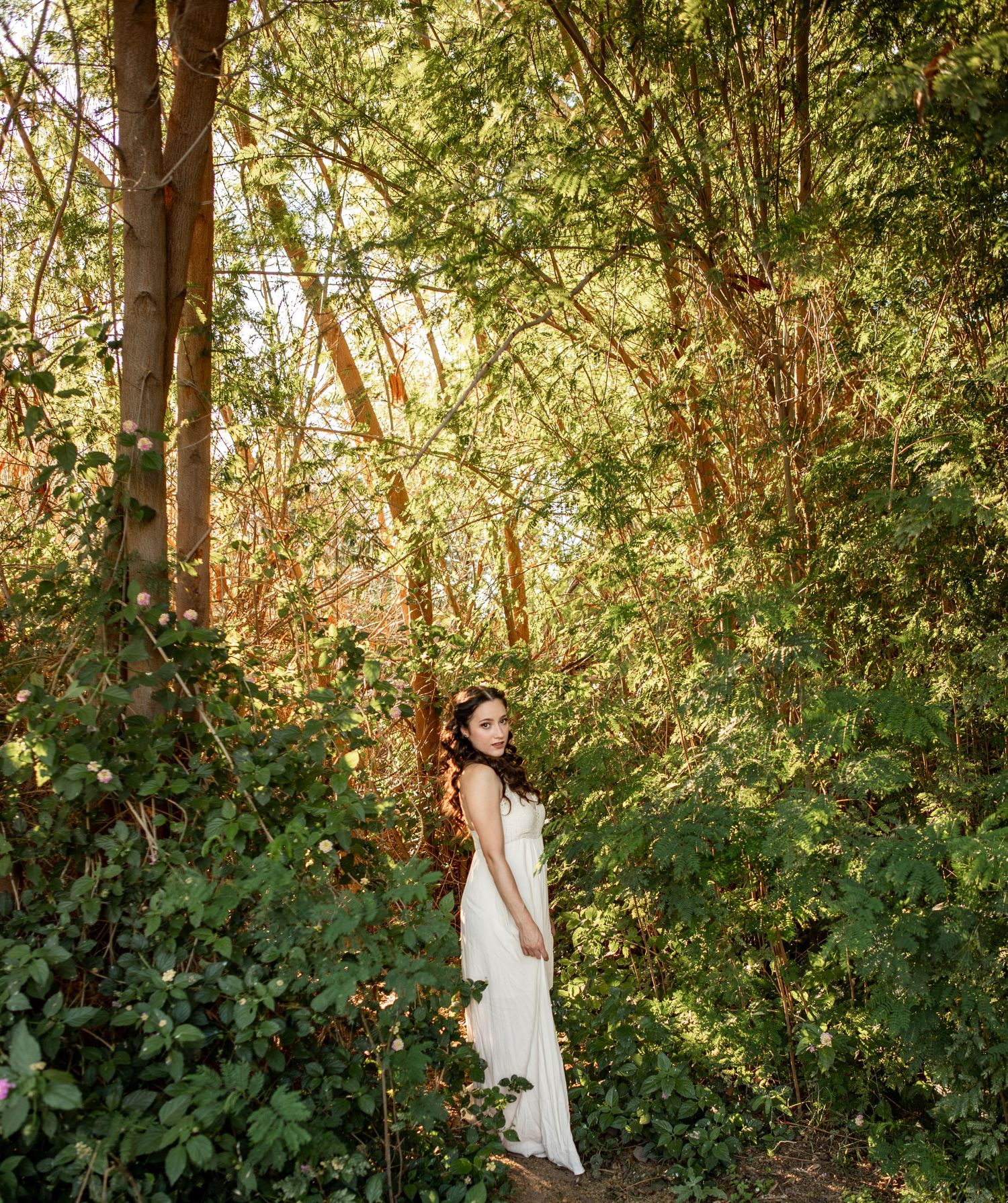 Woman in white dress walking through sunlit forest path surrounded by lush green foliage at The Meadow in Mesa.
