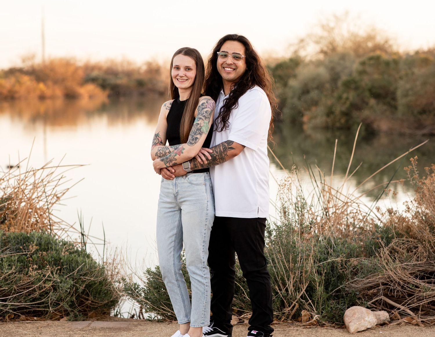 Two friends in casual outfits posing together by a lake during sunset with natural scenery in background.