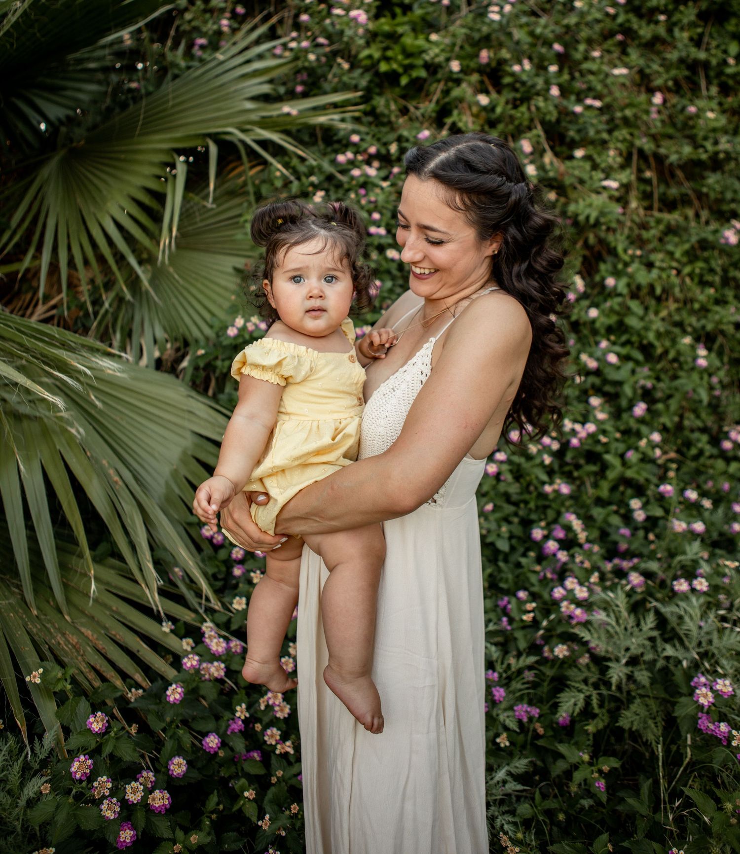 Mother and child in white dress stand among lush greenery and pink flowers in a garden setting at The Meadow in Mesa.