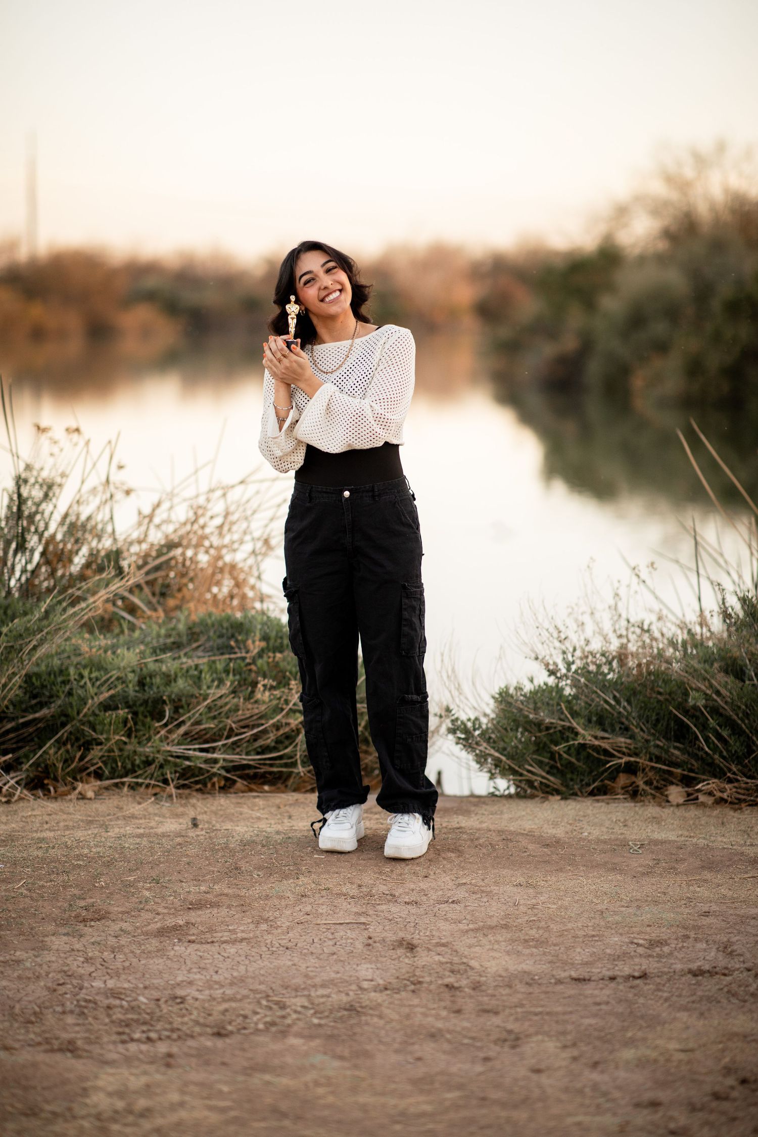 A person wearing black pants and white sneakers sits by a tranquil lake during sunset.