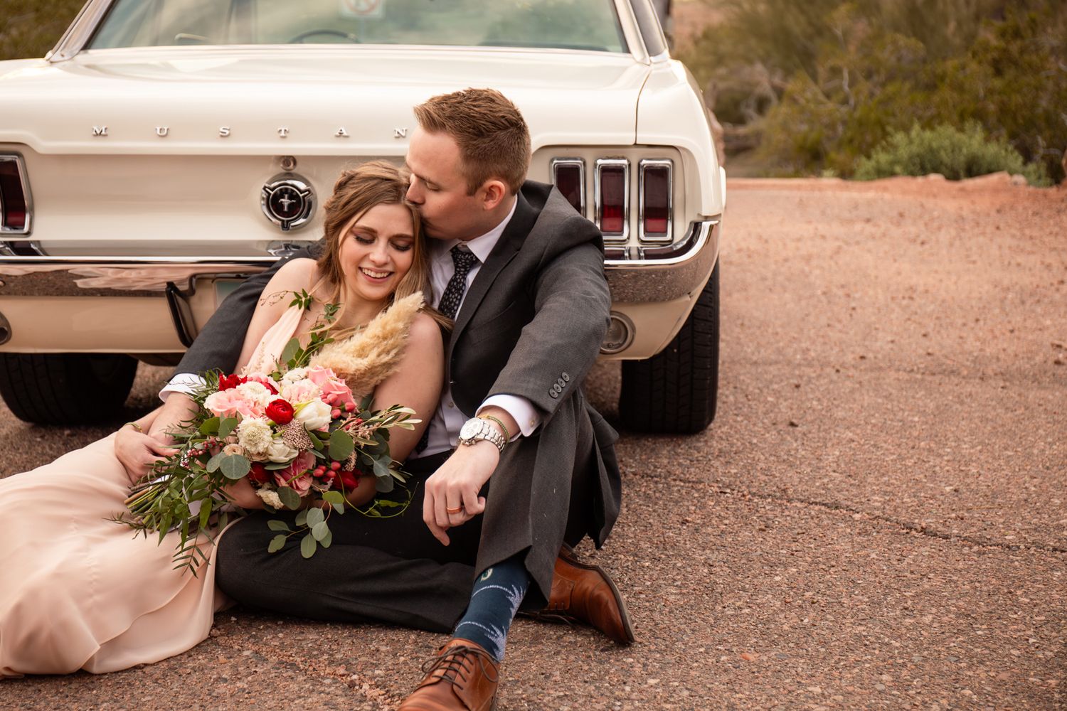 A romantic moment captured in front of a vintage car with red roses and elegant formal attire in Papago Park in Phoenix, AZ.