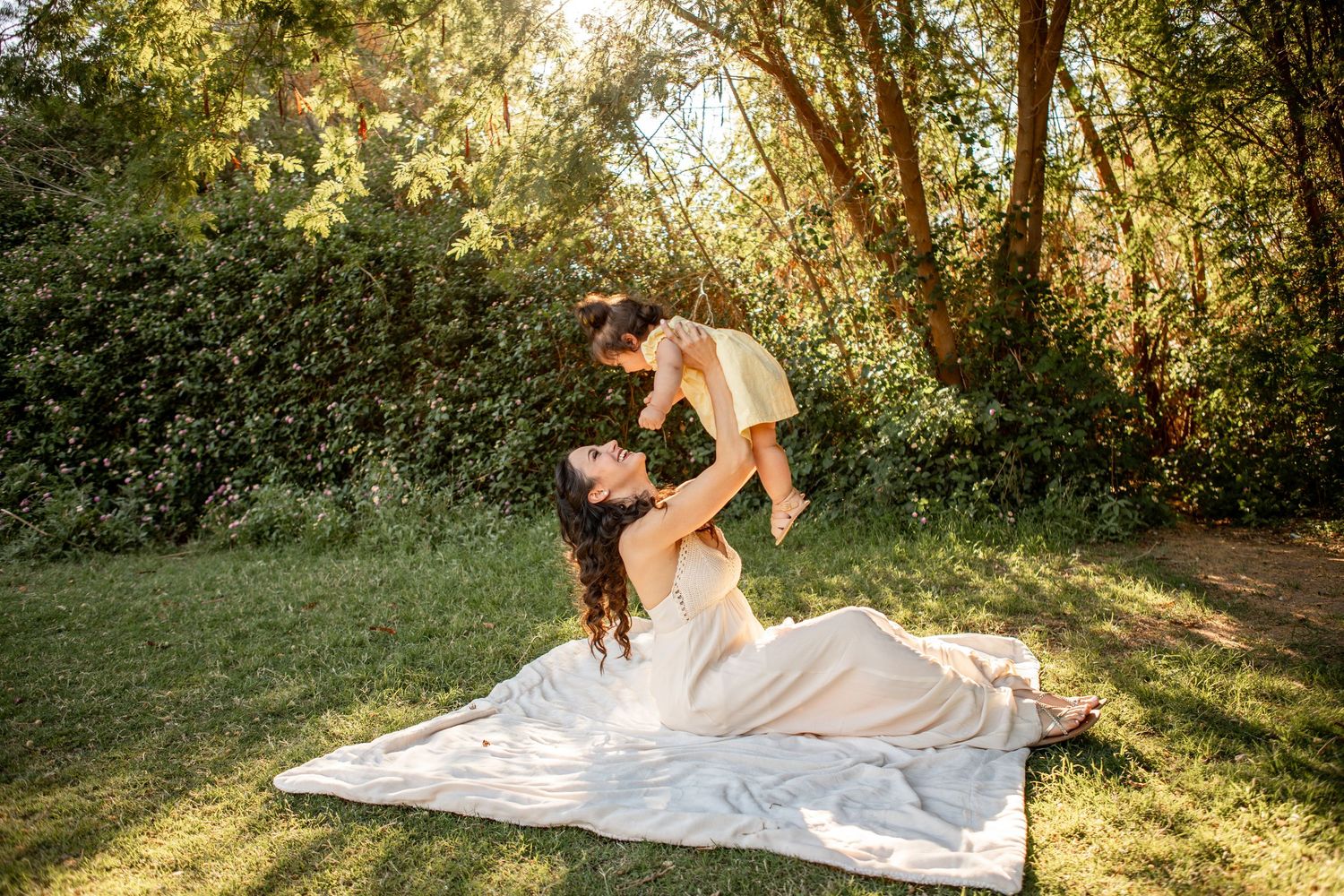 A joyful outdoor scene on a white blanket with sunlit trees and greenery in the background at The Meadow in Mesa.