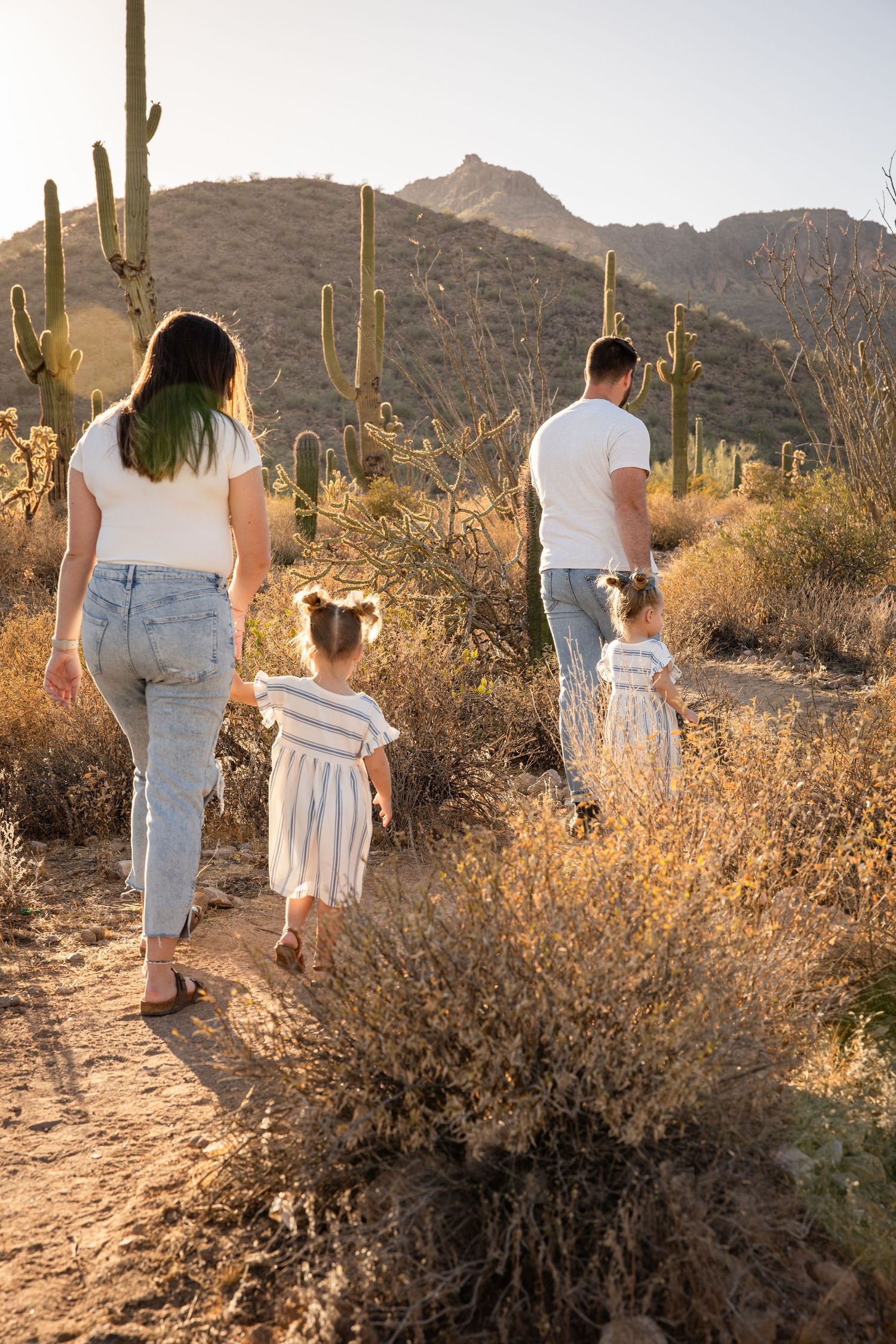 Family of four walking on a desert trail surrounded by saguaro cacti and mountains at sunset.