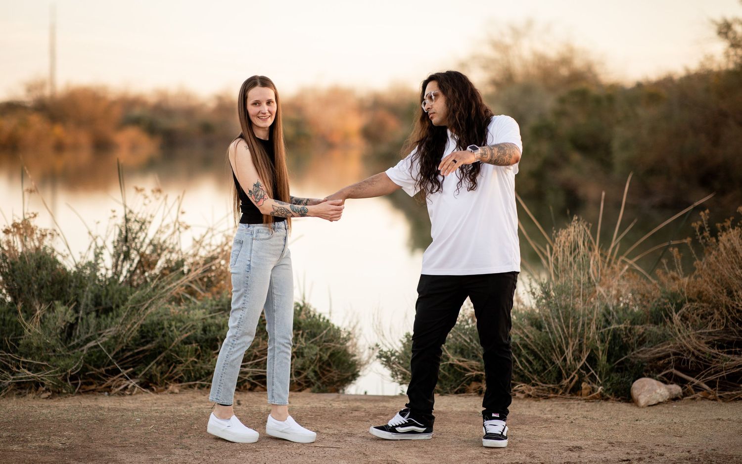 A couple in white and black outfits poses together during golden hour in a desert landscape setting.