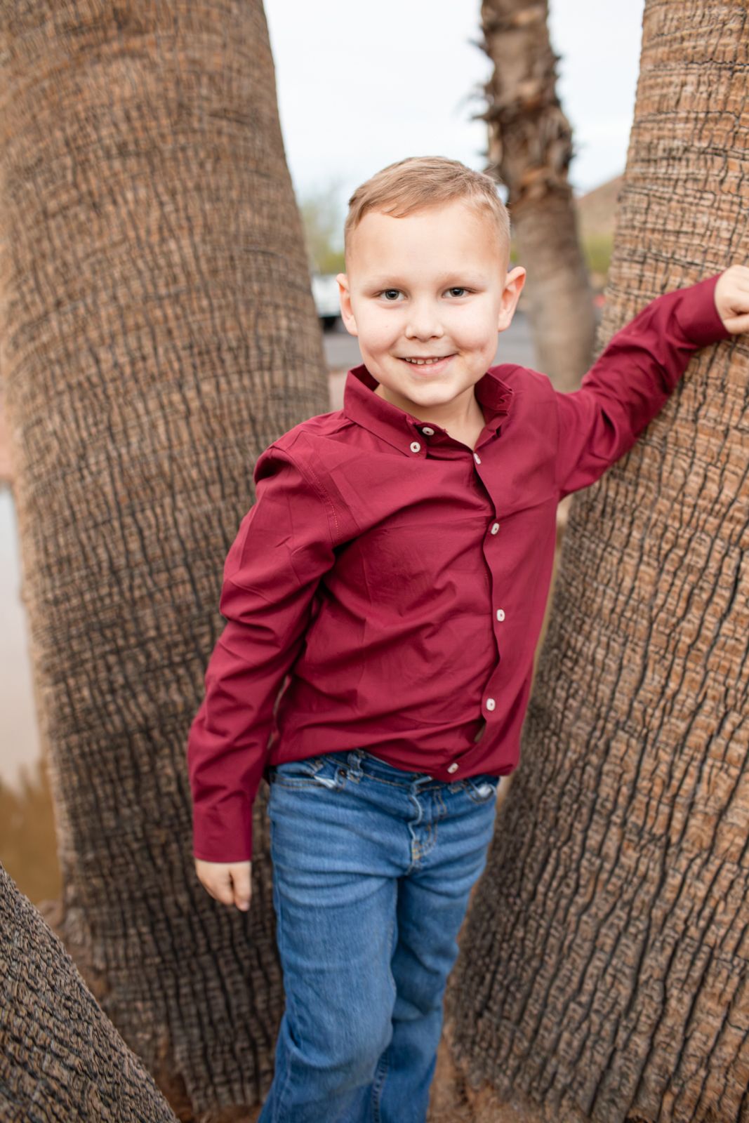 A young child in a burgundy shirt and jeans poses playfully between palm tree trunks in an outdoor setting in Papago Park in Phoenix, AZ.