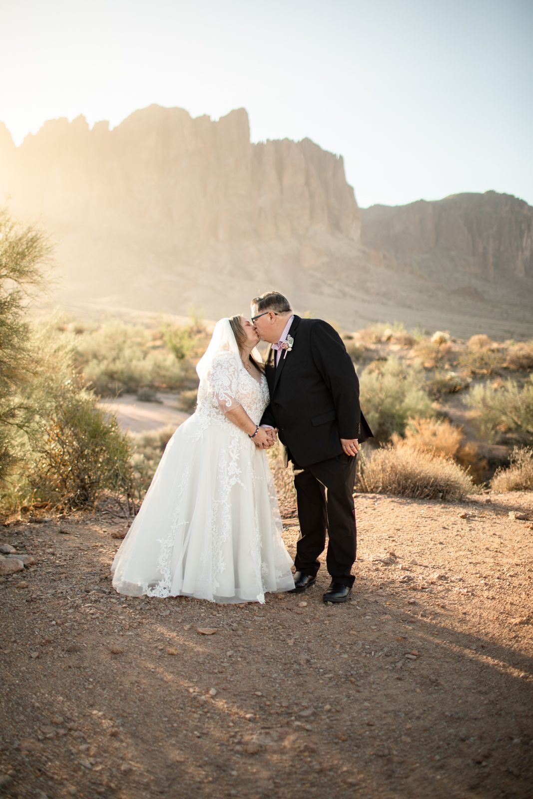 A couple in wedding attire share a romantic moment against the superstition mountains backdrop at sunset.