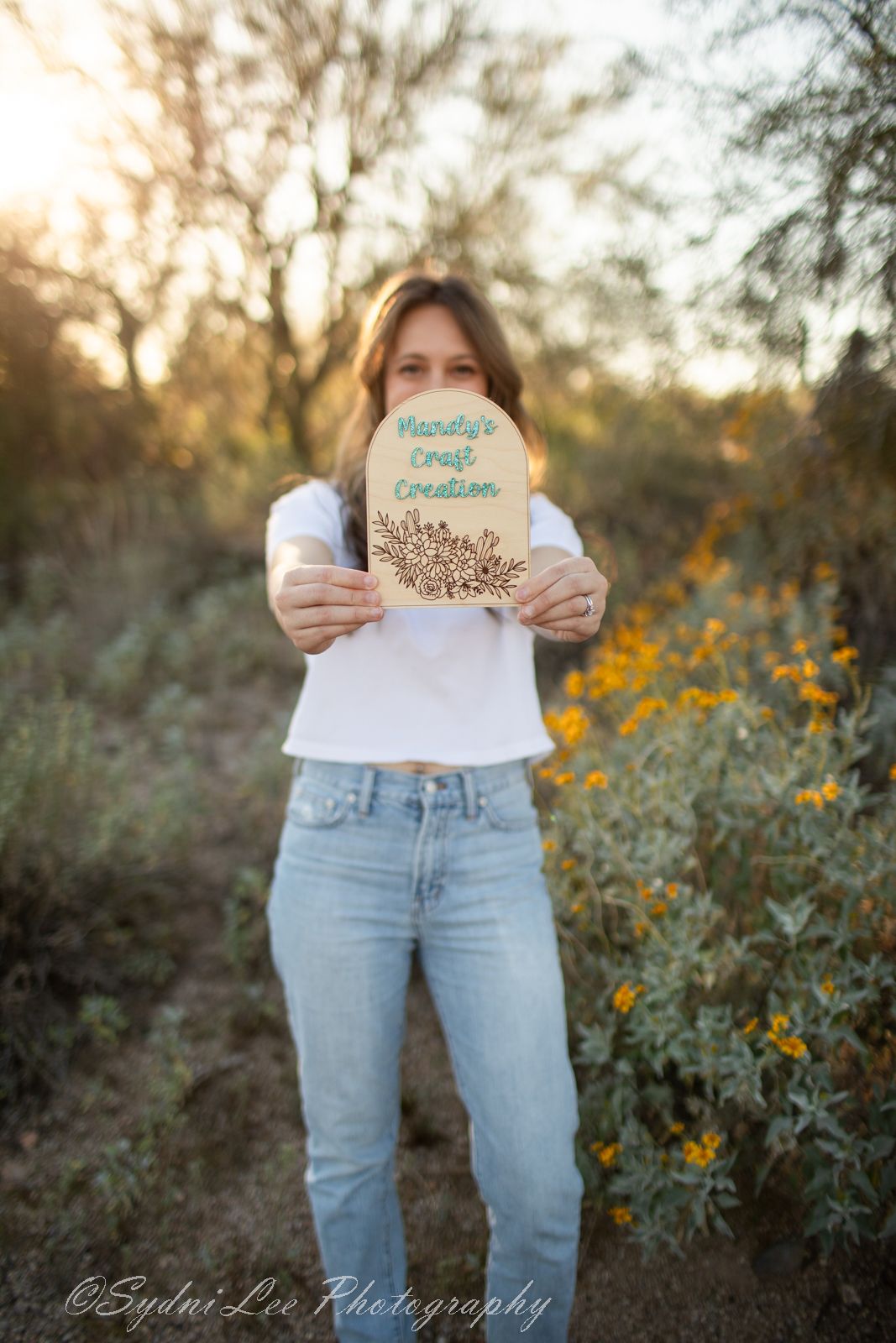 Person in light jeans holds up a small circular wooden sign during golden hour in a desert garden setting.