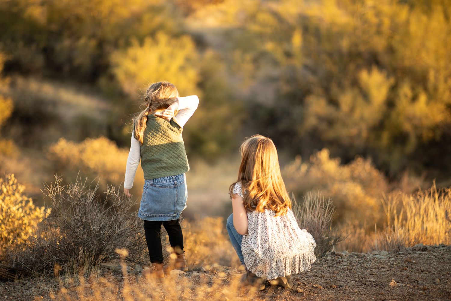 Two young children in white dresses walk along a desert trail during golden hour as sunlight illuminates the landscape at Lost Dutchman
