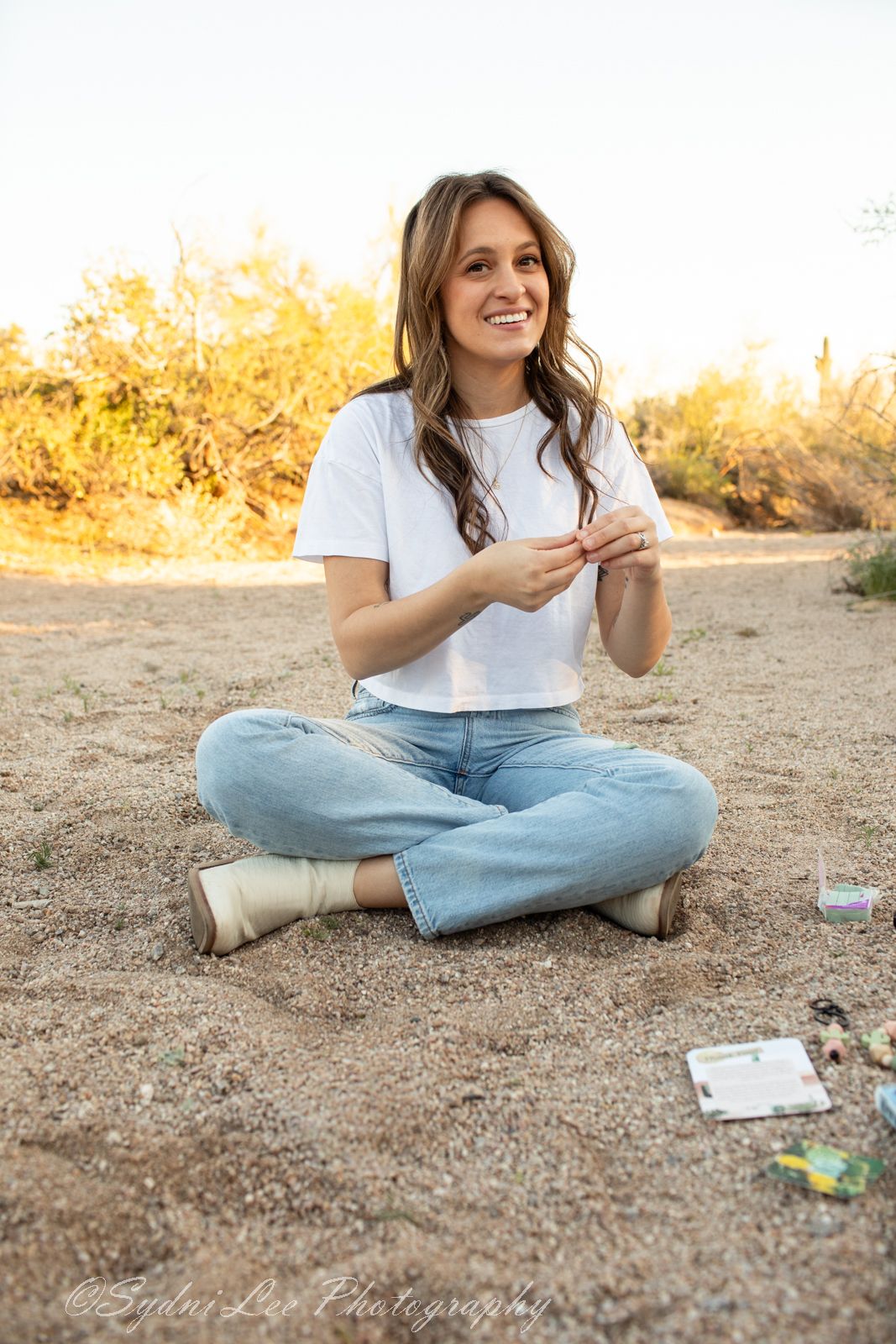Person in casual attire sitting cross-legged on desert ground during golden hour.