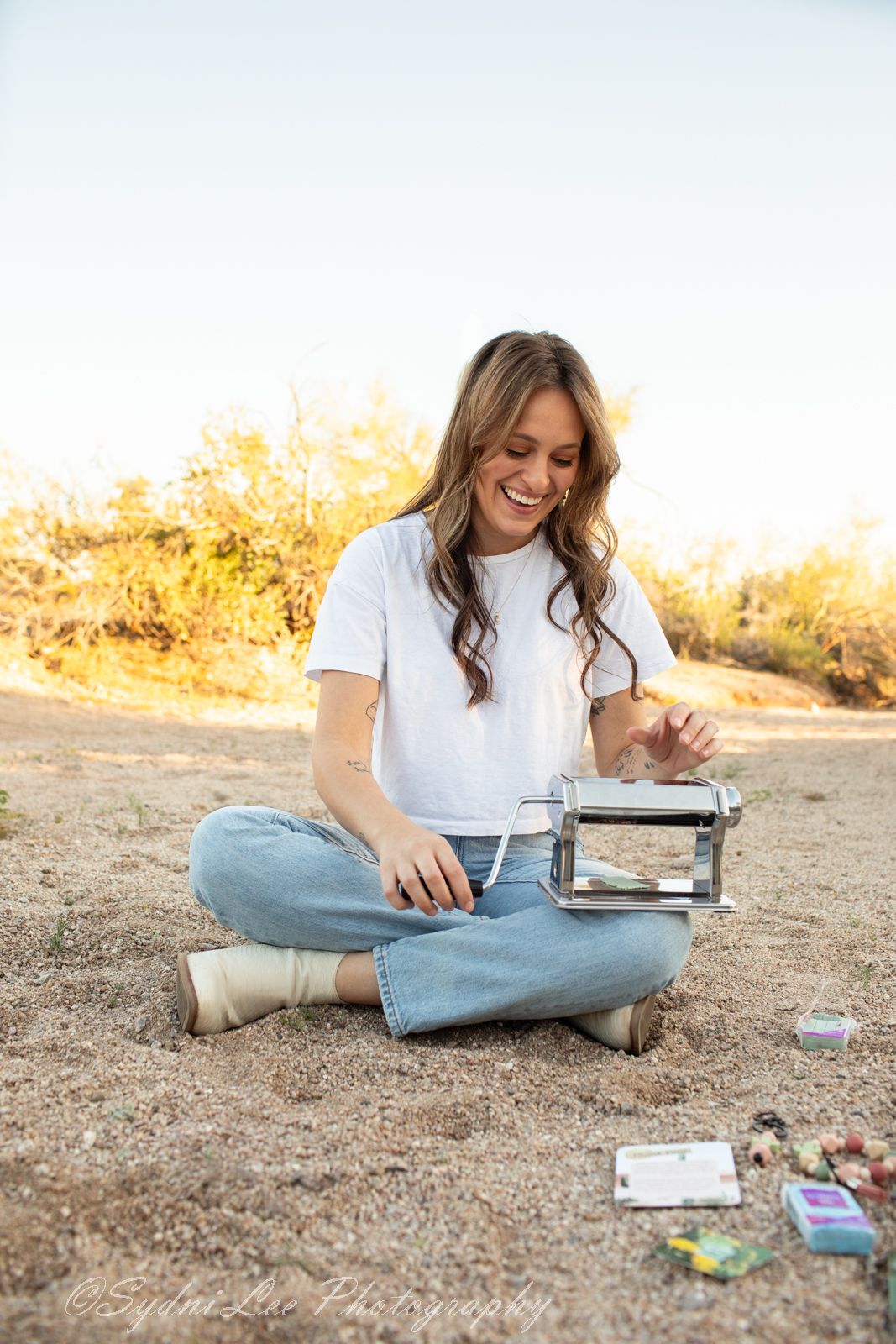 Person in white t-shirt and jeans sits cross-legged on desert ground writing in journal at golden hour.