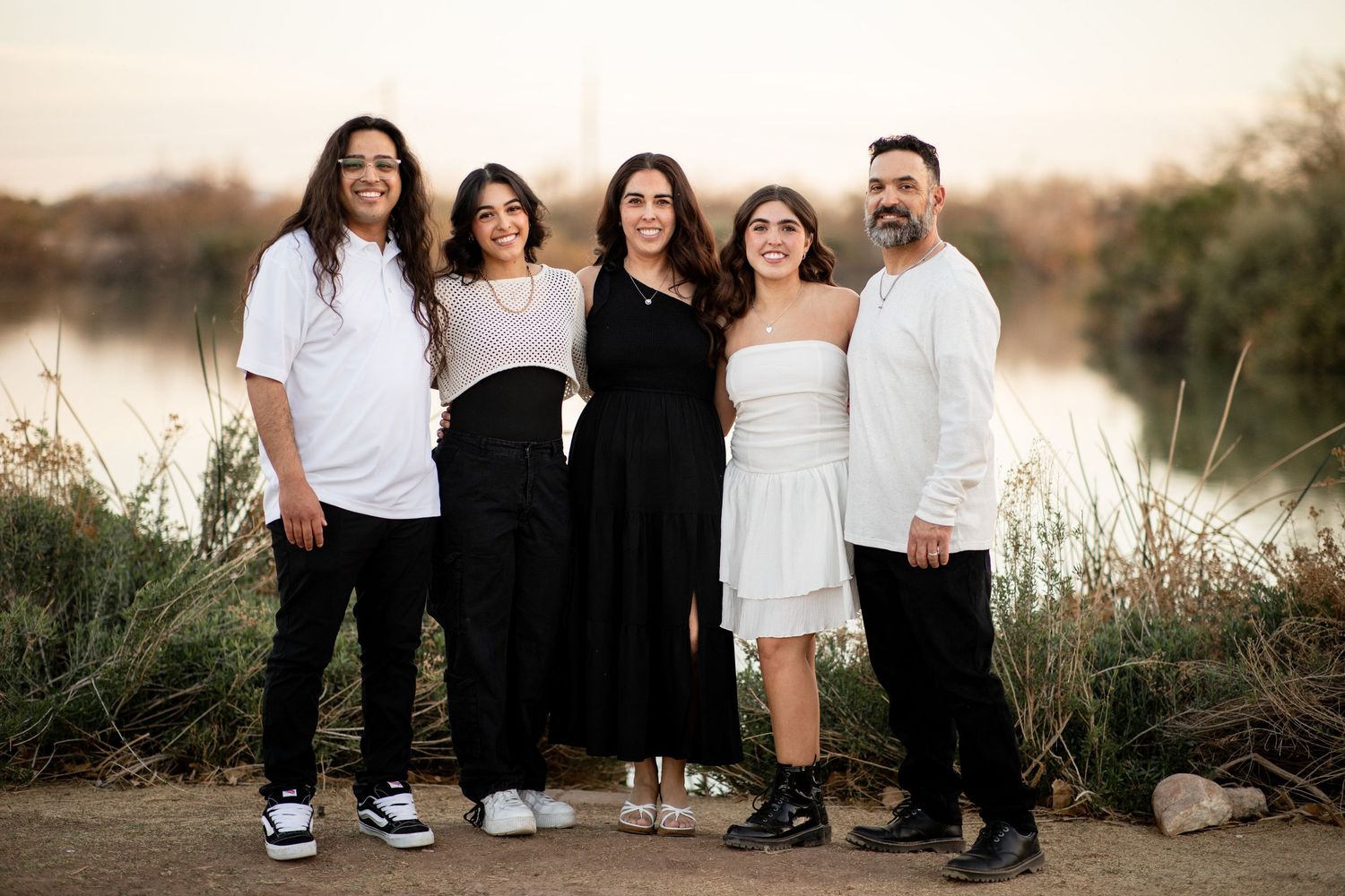 A group of five people in coordinated black and white outfits pose together outdoors at sunset near a lake.