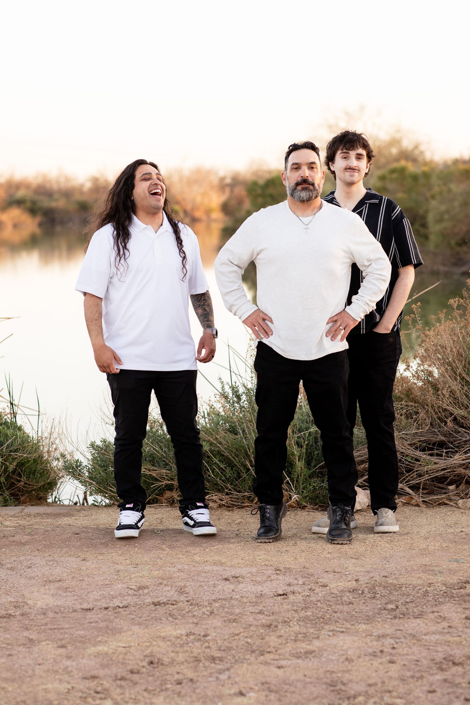 Three people standing together and laughing on a dirt path by a lake during golden hour.