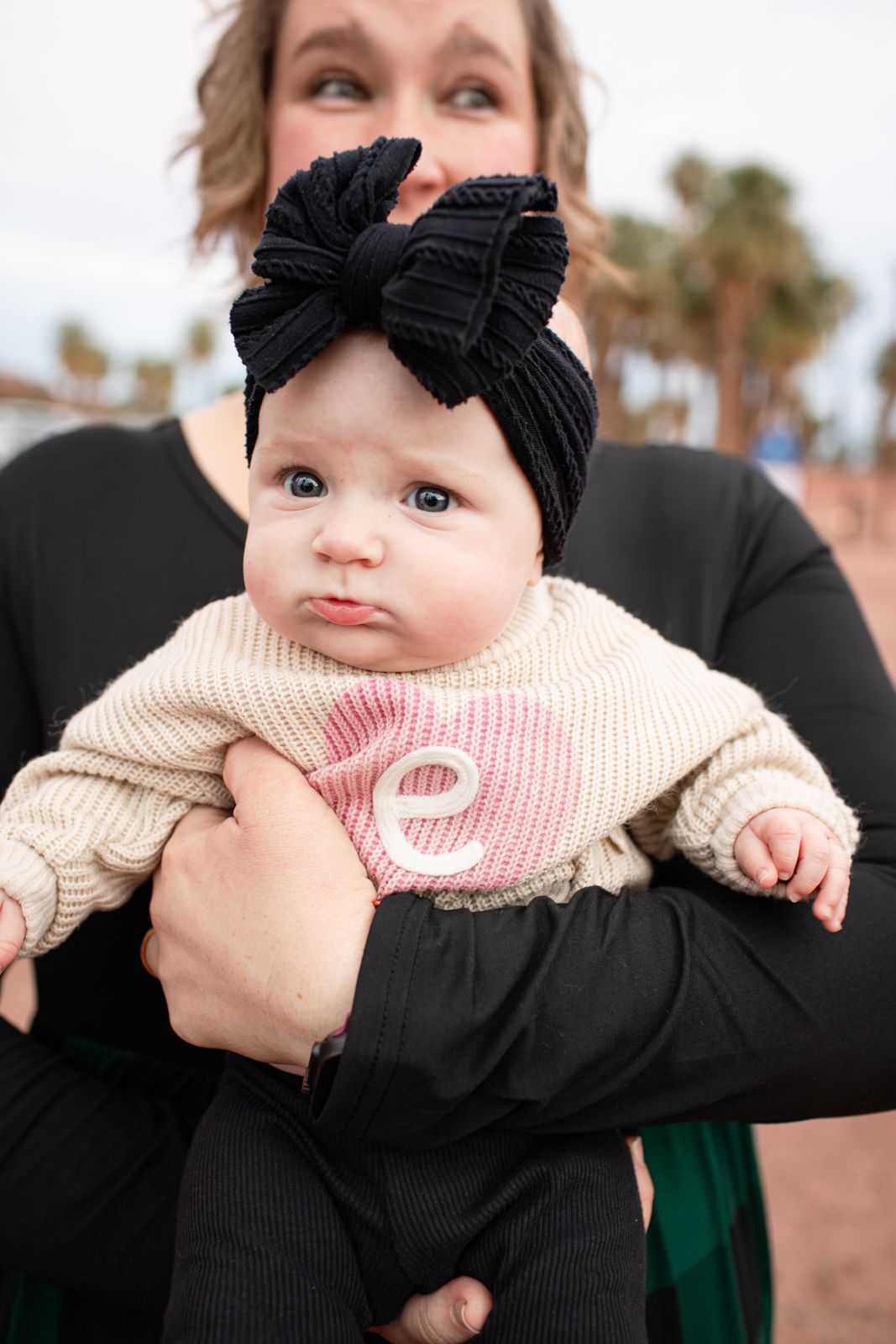 A close-up portrait of a baby in a cream sweater and black bow headband being held in Papago Park in Phoenix, AZ.