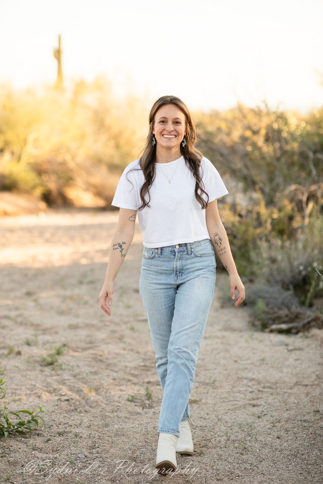 A person in a white t-shirt and light jeans walks along a desert trail during golden hour sunset.
