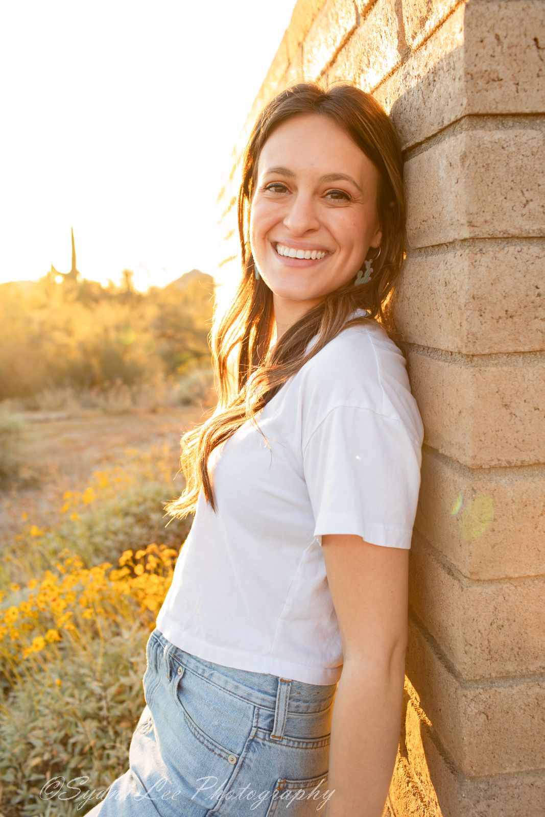 Person in white t-shirt and jeans poses against stone wall during golden hour sunset in desert landscape.