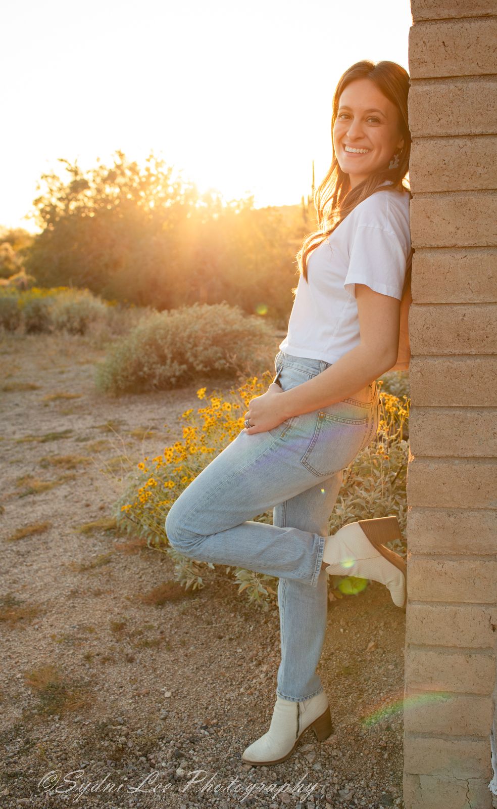 Person in white t-shirt and jeans leaning against brick wall at sunset with golden sunlight and desert brush in background.
