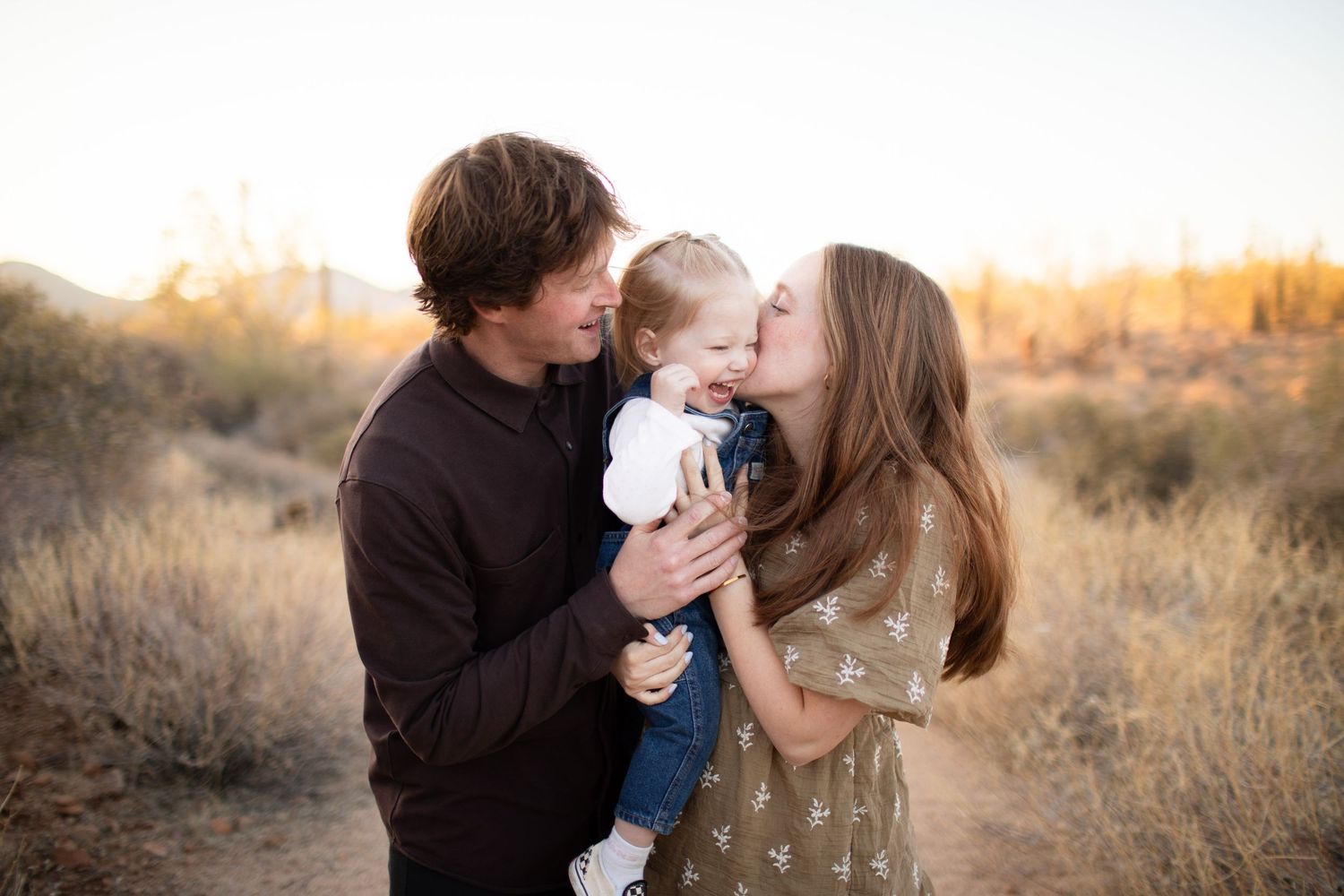 A young family shares a sweet moment outdoors at sunset, parents kissing their toddler on a desert trail.