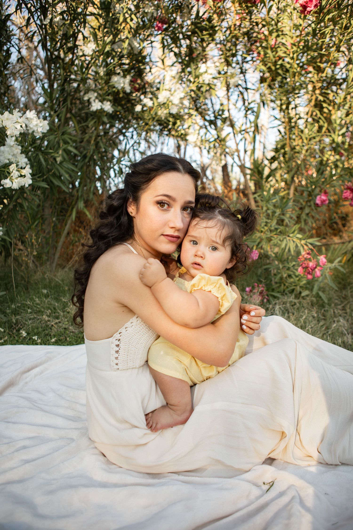 A tender outdoor photoshoot featuring a parent and child in light-colored dresses laying on a white blanket at The Meadow in Mesa.