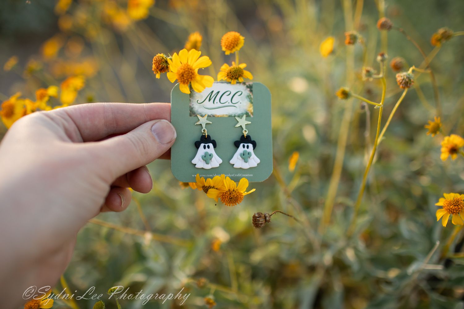 Hand holds small green enamel pin against backdrop of blooming yellow wildflowers.