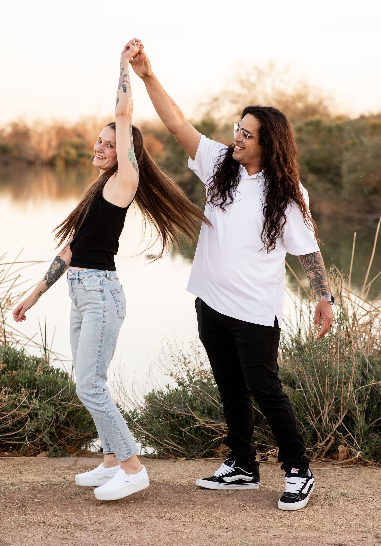Two friends in casual outfits dance and hold hands by a lake at sunset, showing joyful expressions and carefree movement.