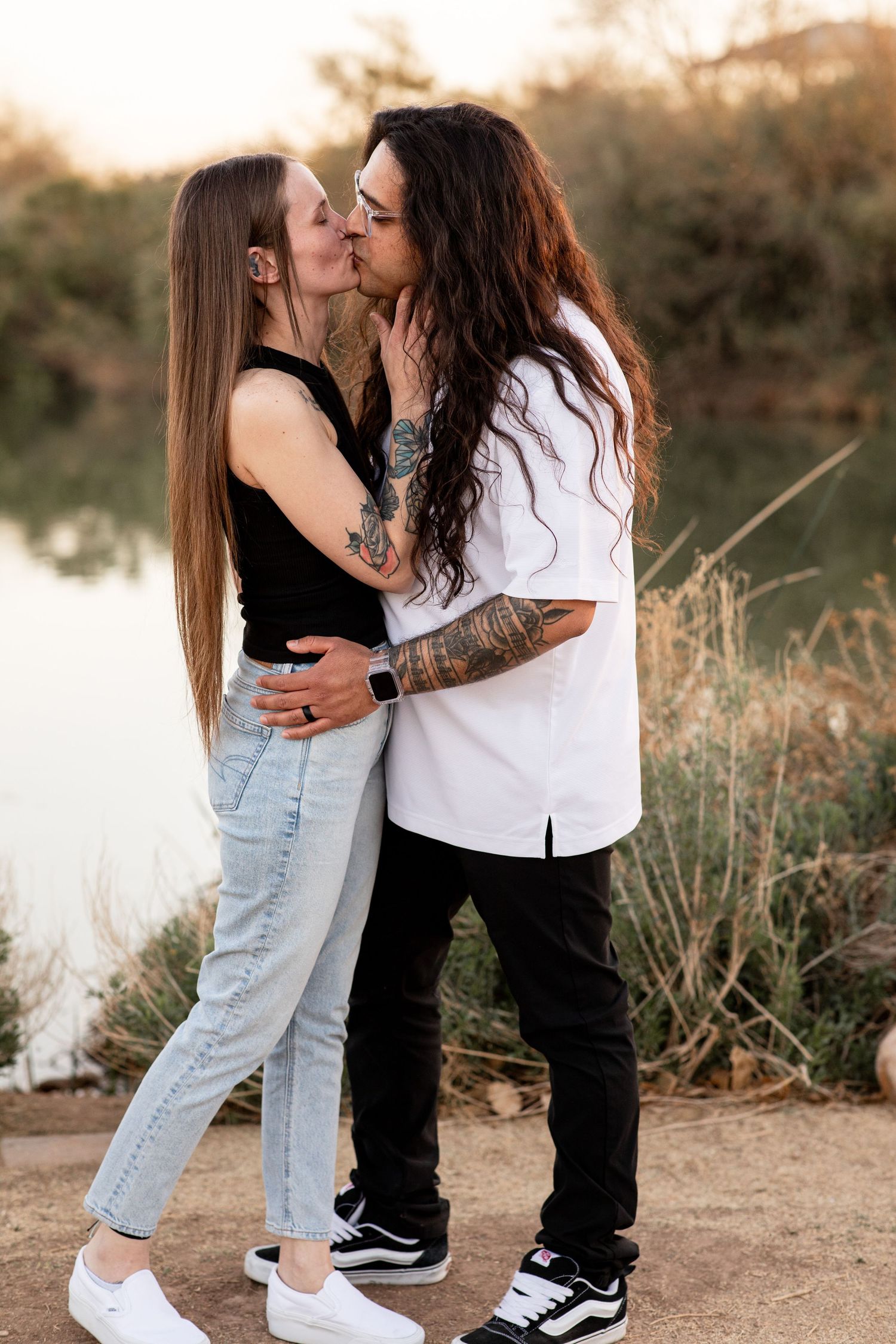 A couple shares an intimate moment in casual attire while standing outdoors at sunset in a natural setting.