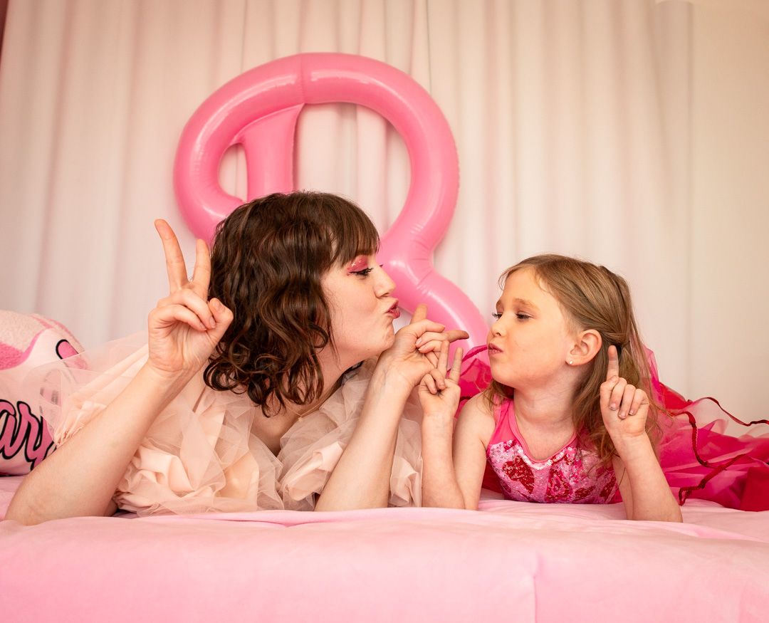 Two people lying on pink bedding making silly faces with pink balloon decoration.