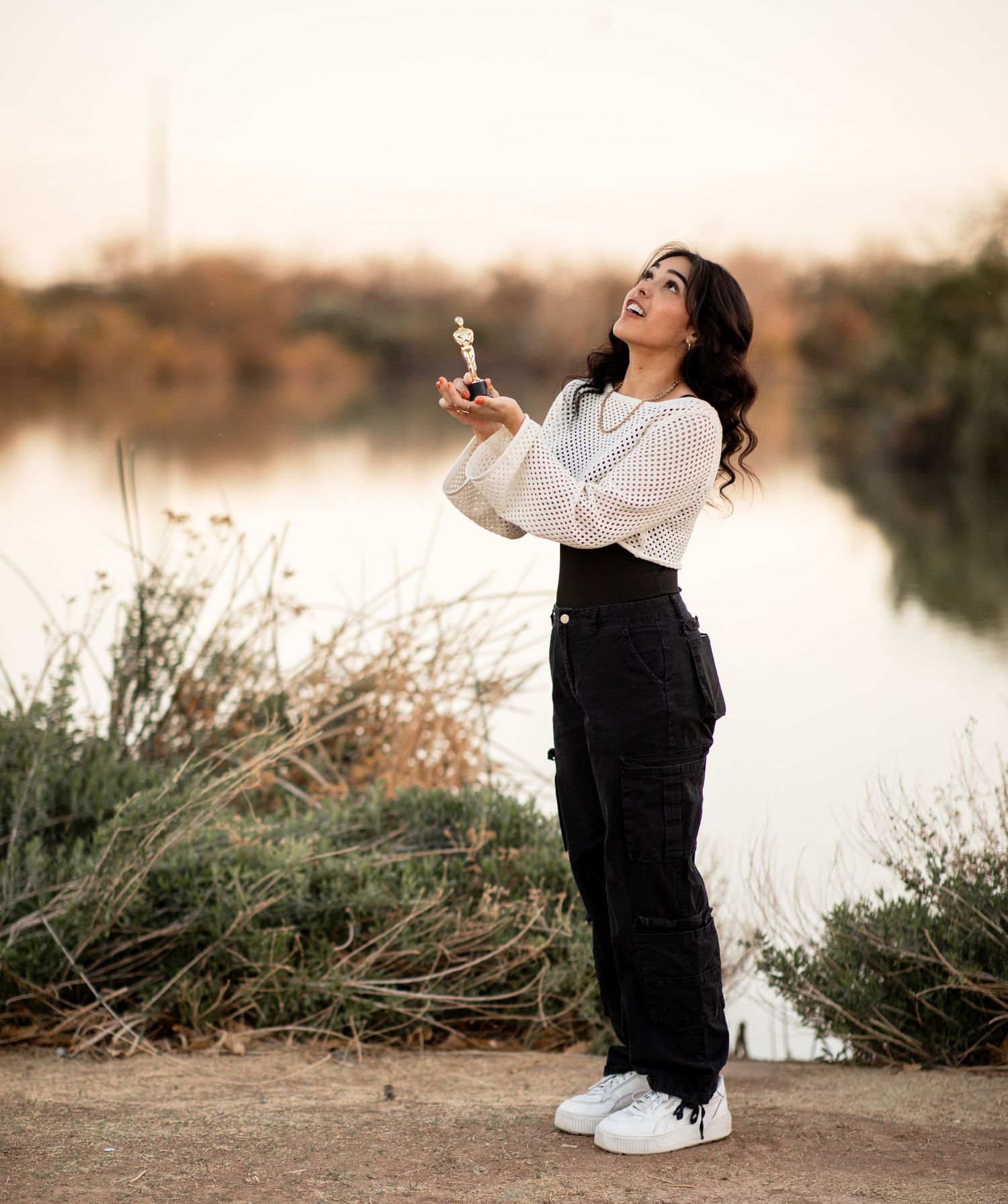 Person in white blouse and black pants poses with dandelion by serene waterfront at sunset.