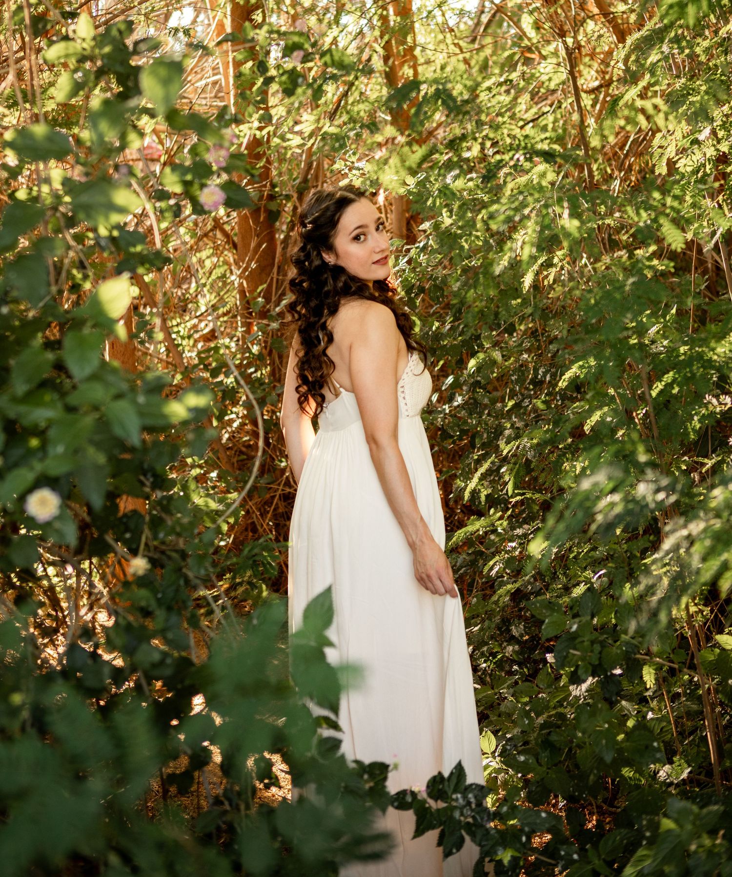 A figure in a flowing white dress poses gracefully among lush green foliage and blooming flowers in a garden setting at The Meadow in Mesa.