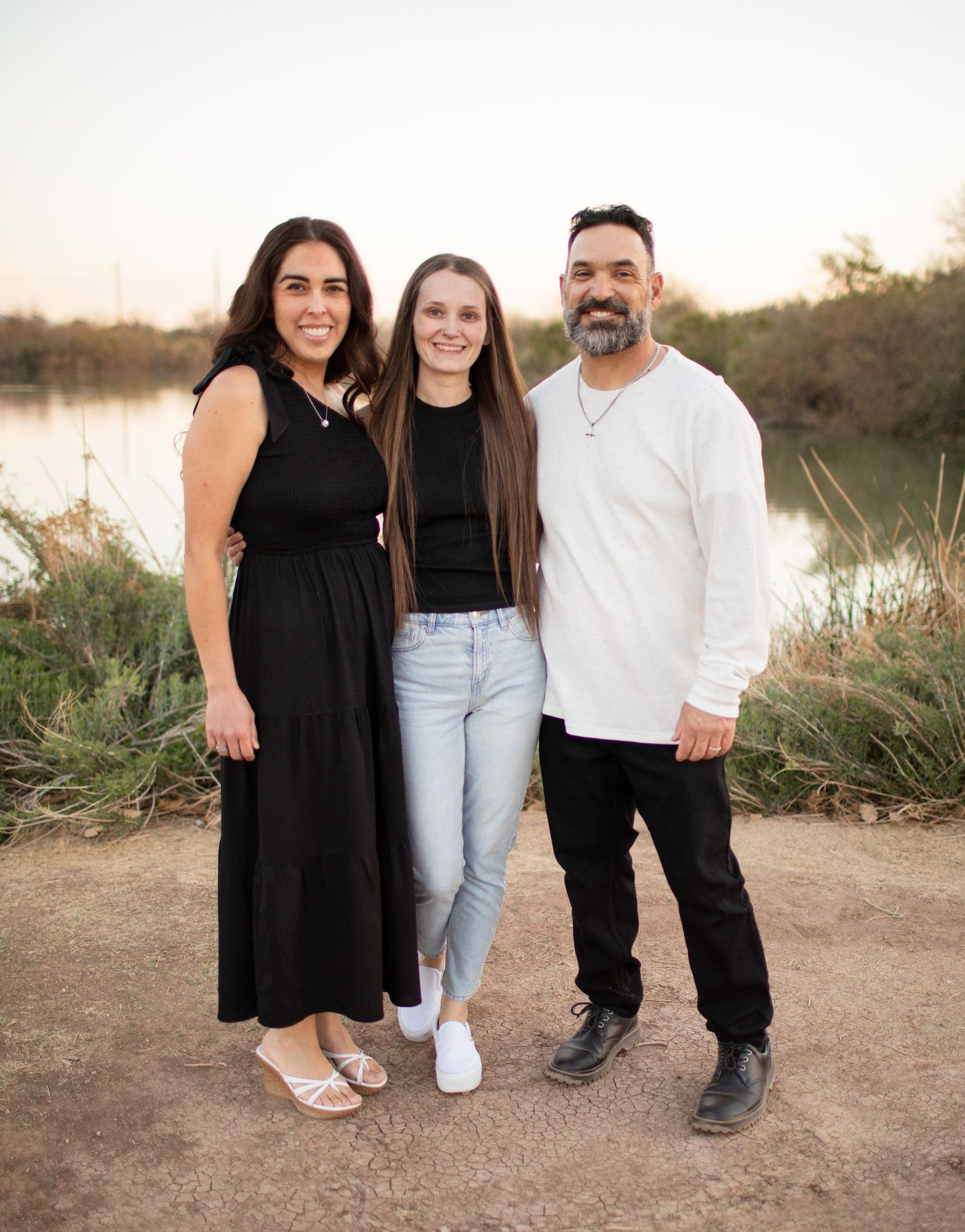Three people stand together on a dirt path near a lake at sunset in a casual outdoor portrait setting.
