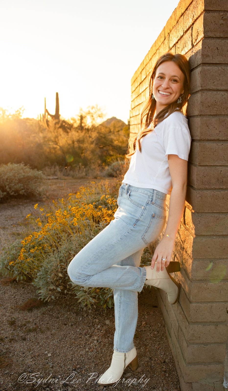 Person in light jeans and white top poses against stone wall during golden desert sunset with yellow wildflowers.