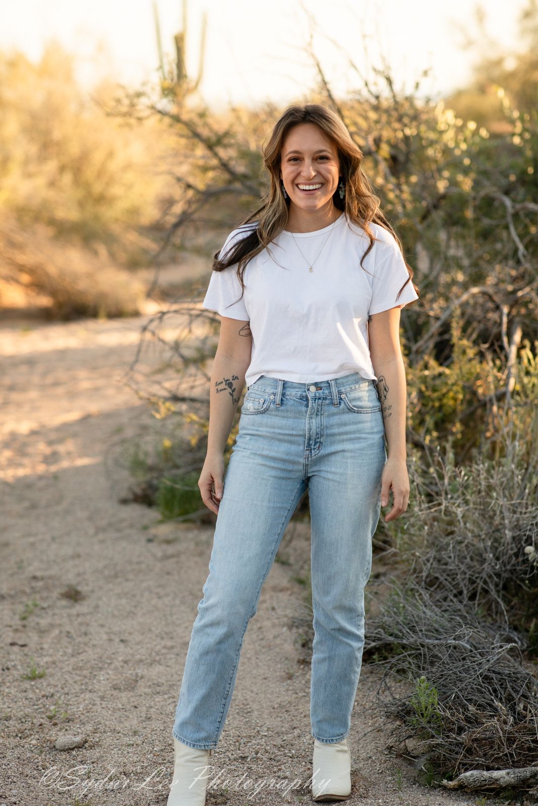 Person in white t-shirt and light denim stands on desert trail during sunset.