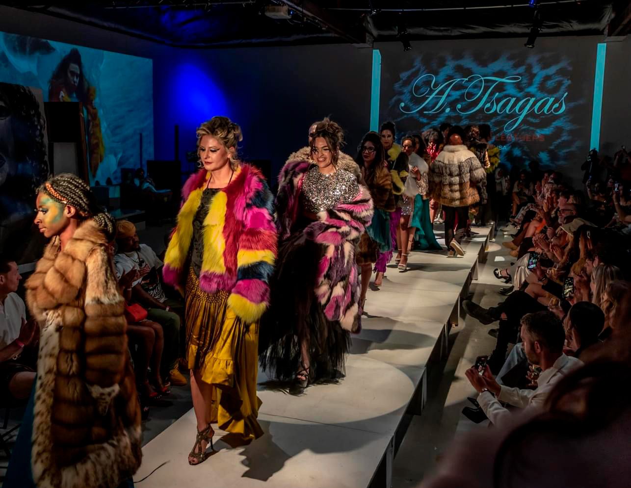 Fashion show runway event with blue lighting and audience members seated along sides.