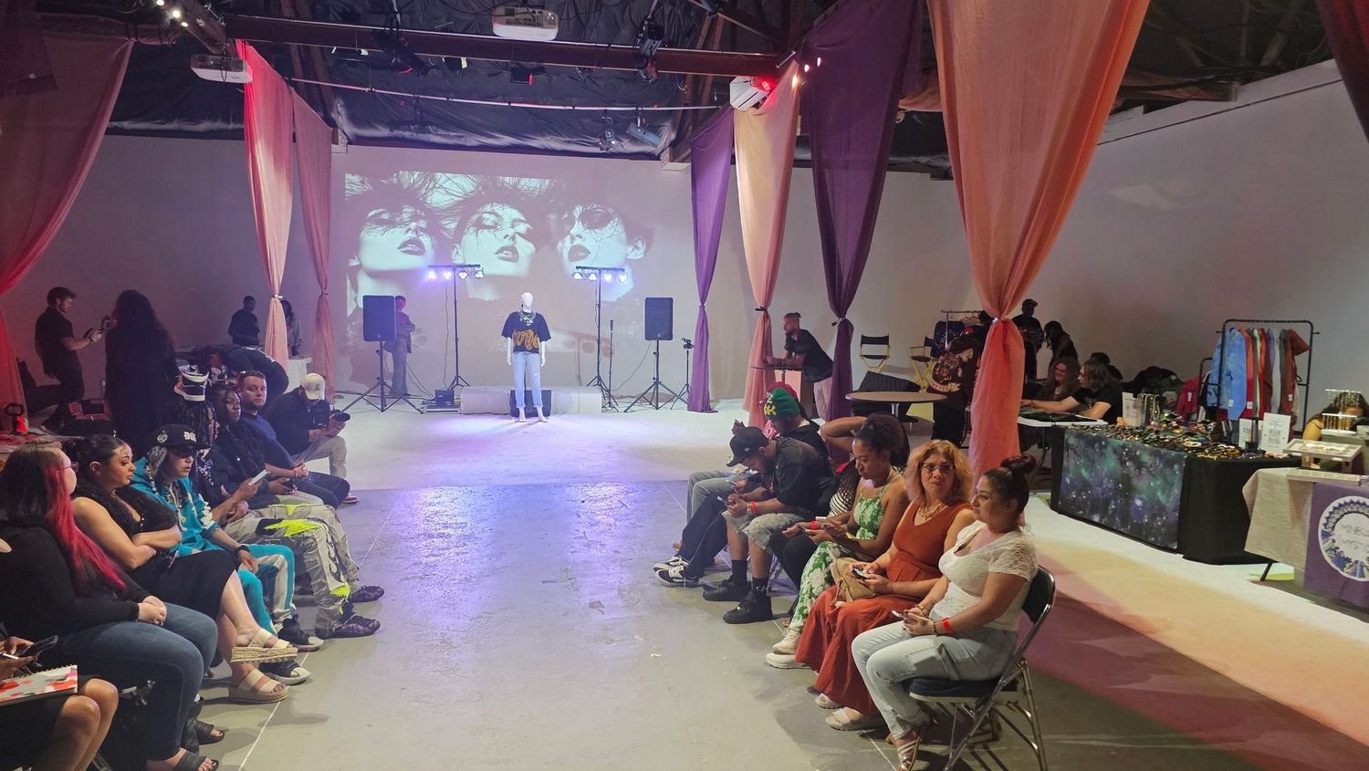An event space with people seated along both sides watching a staged performance area with pink draping and purple lighting.