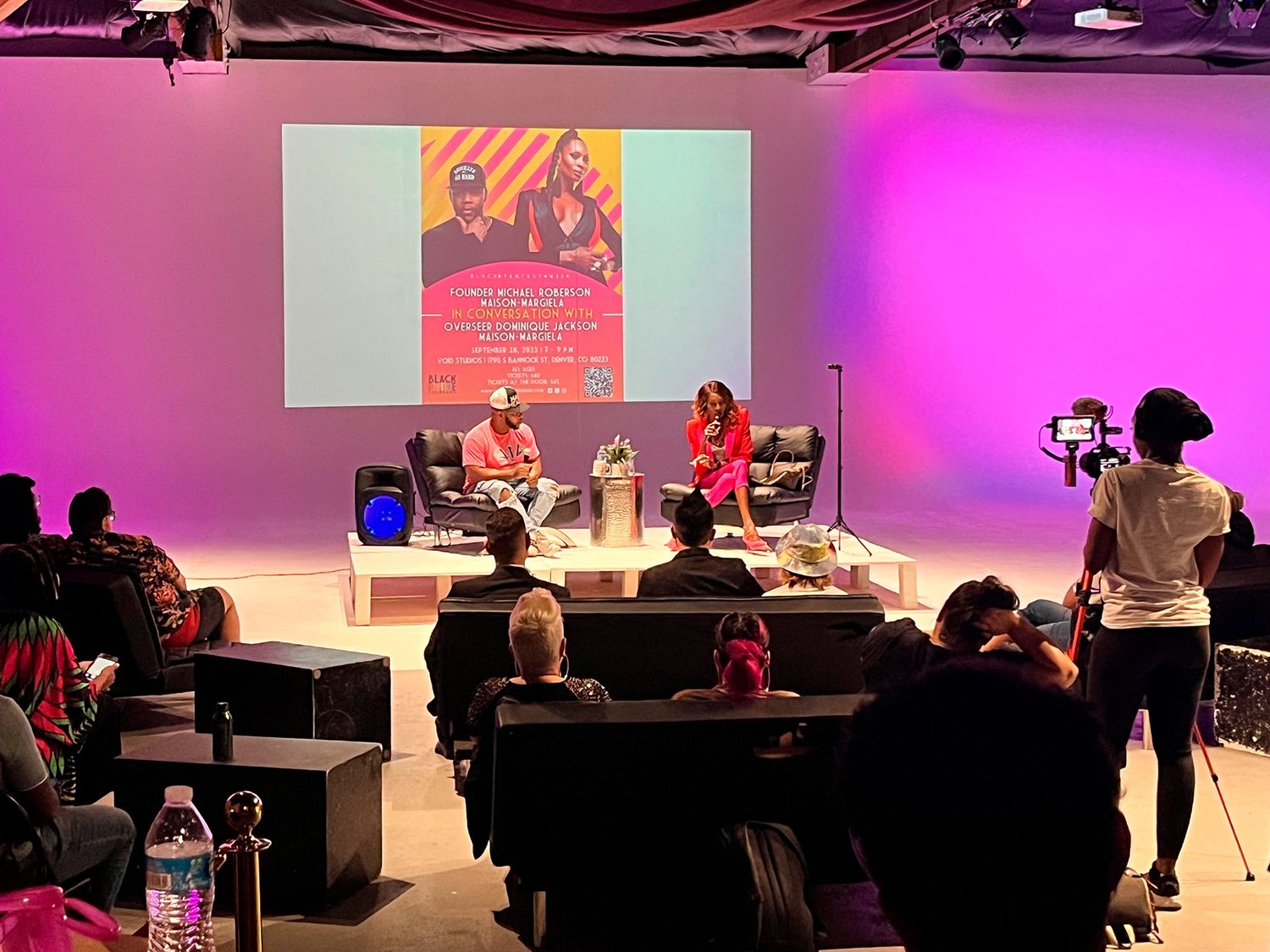 Conference presentation event showing audience members seated in rows watching speakers on a colorful lit stage.