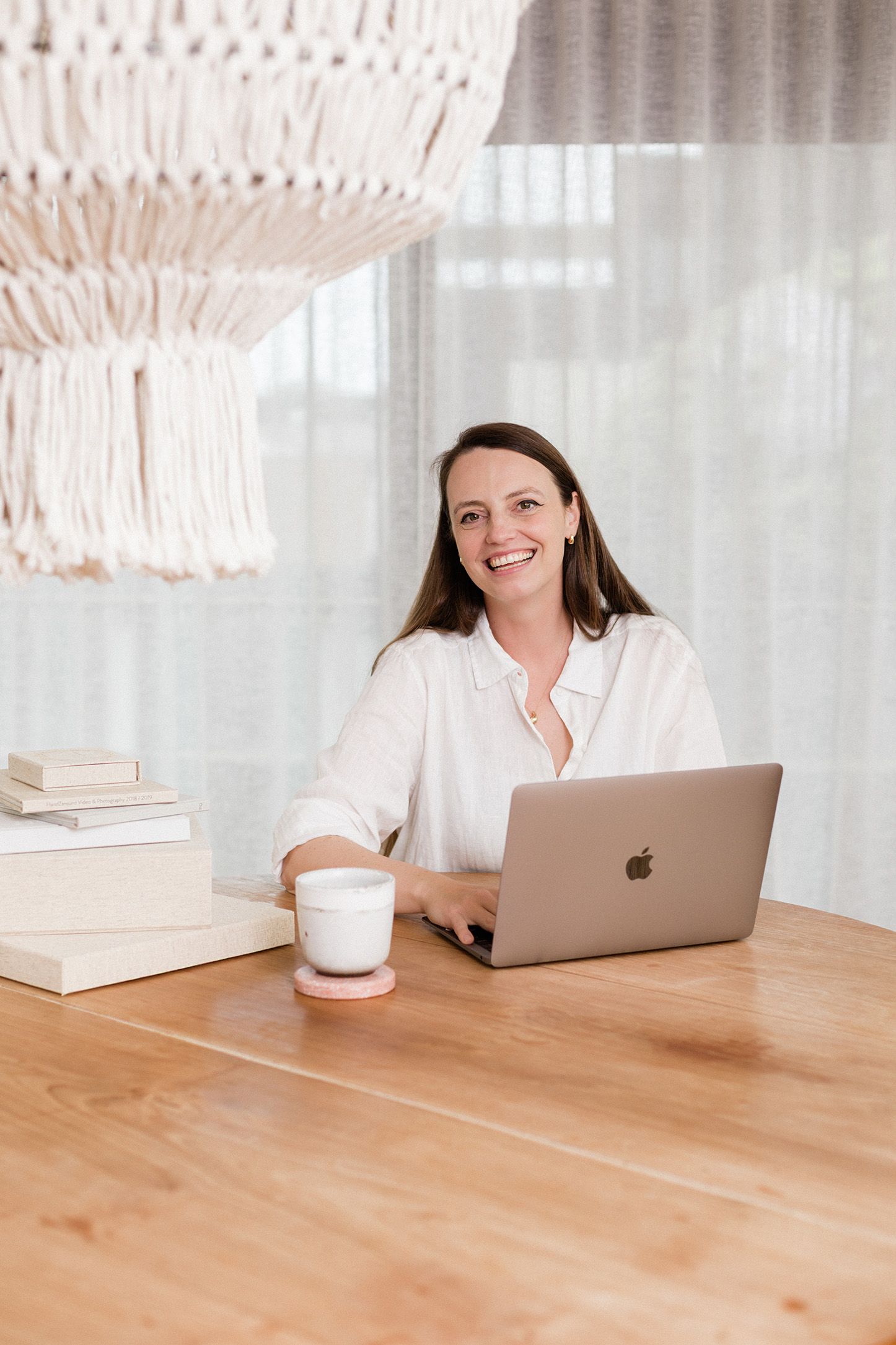 Woman smiling at desk with MacBook laptop, coffee cup, and books in bright home office setting.