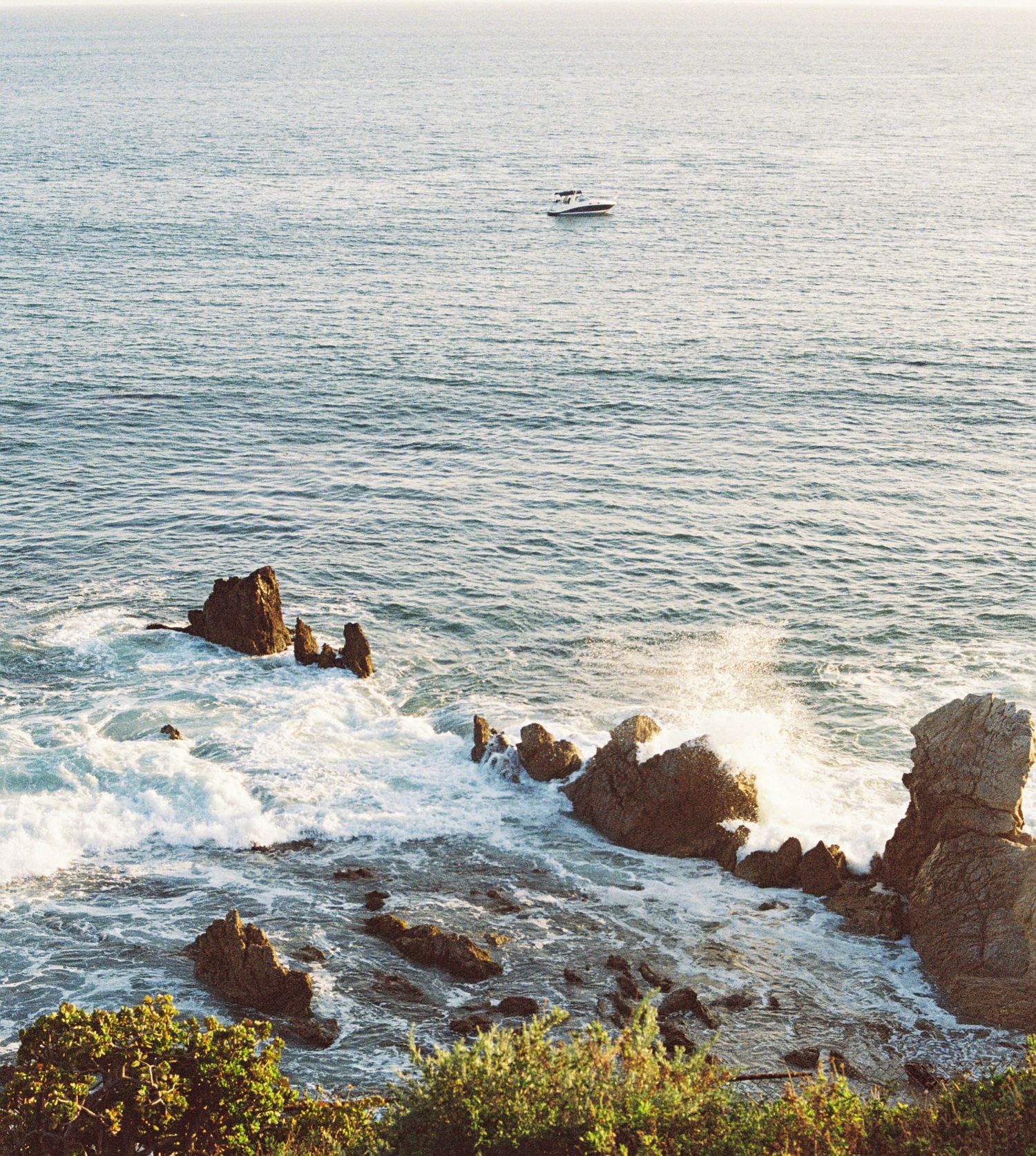 Scenic coastal view of rocky shoreline with crashing waves and small boat in distance.