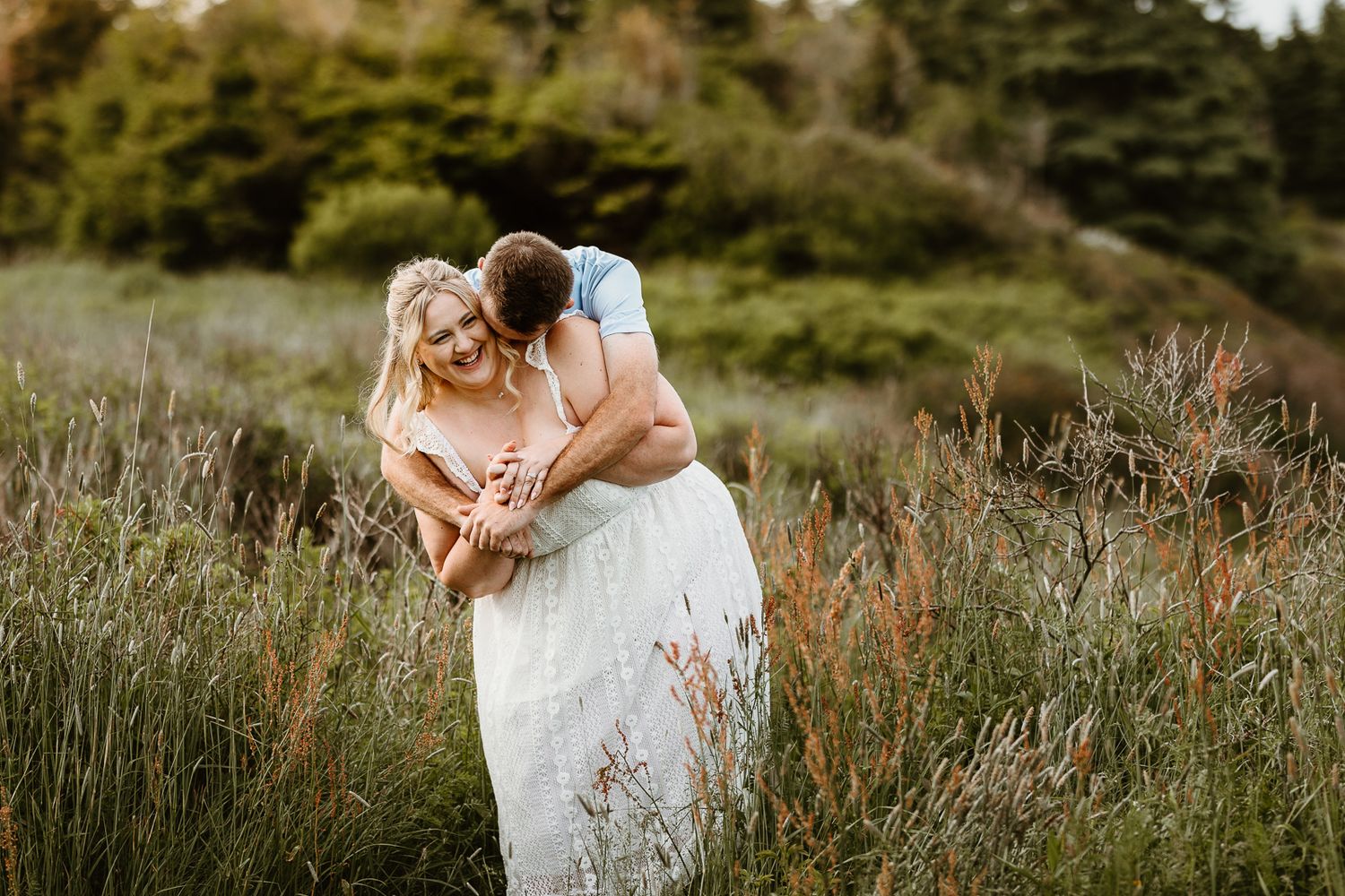 Couple embracing in tall grass field, bride in white gown laughing as groom hugs her from behind at golden hour.