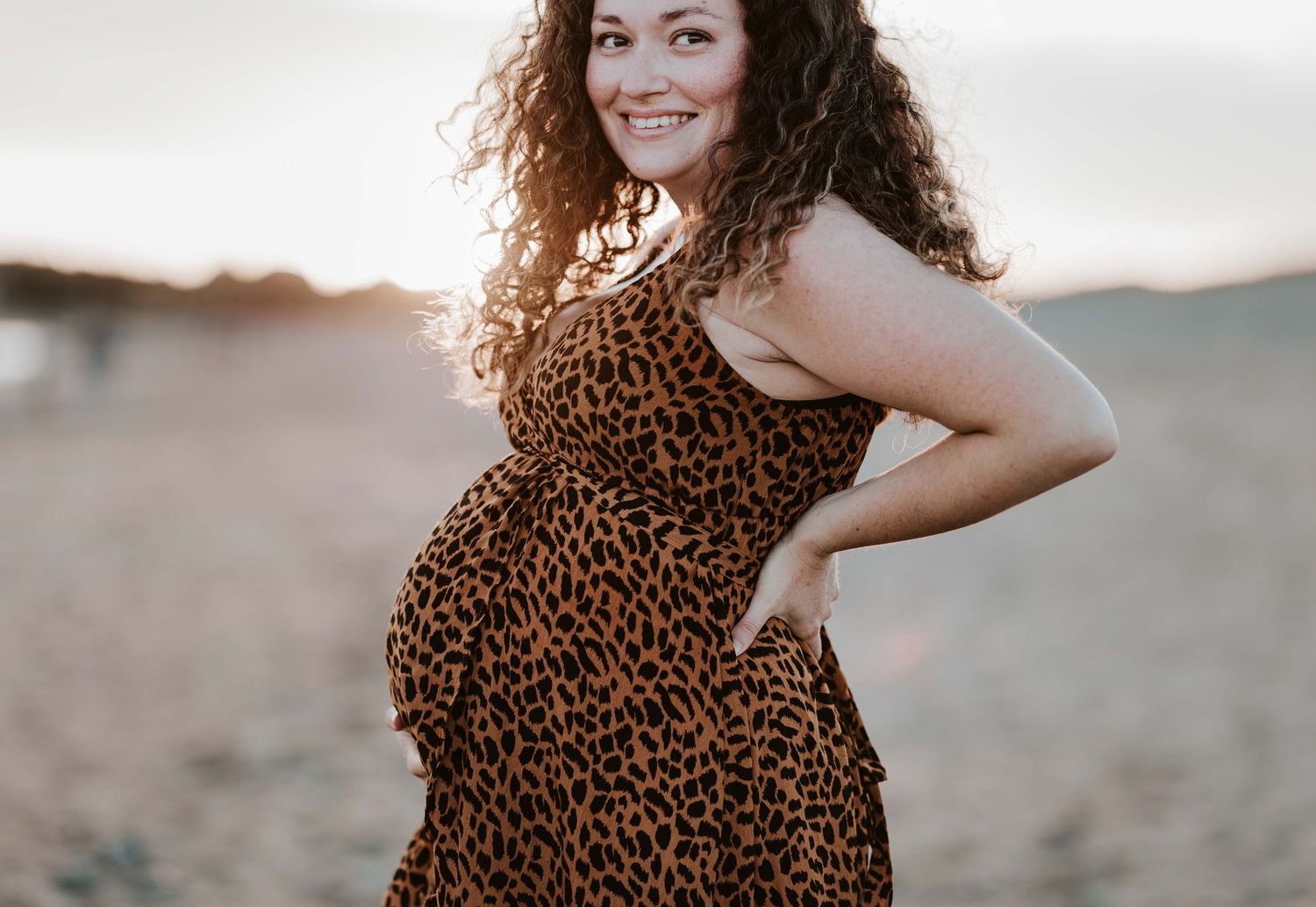Maternity photoshoot on beach features curly-haired figure in leopard print dress against ocean backdrop.